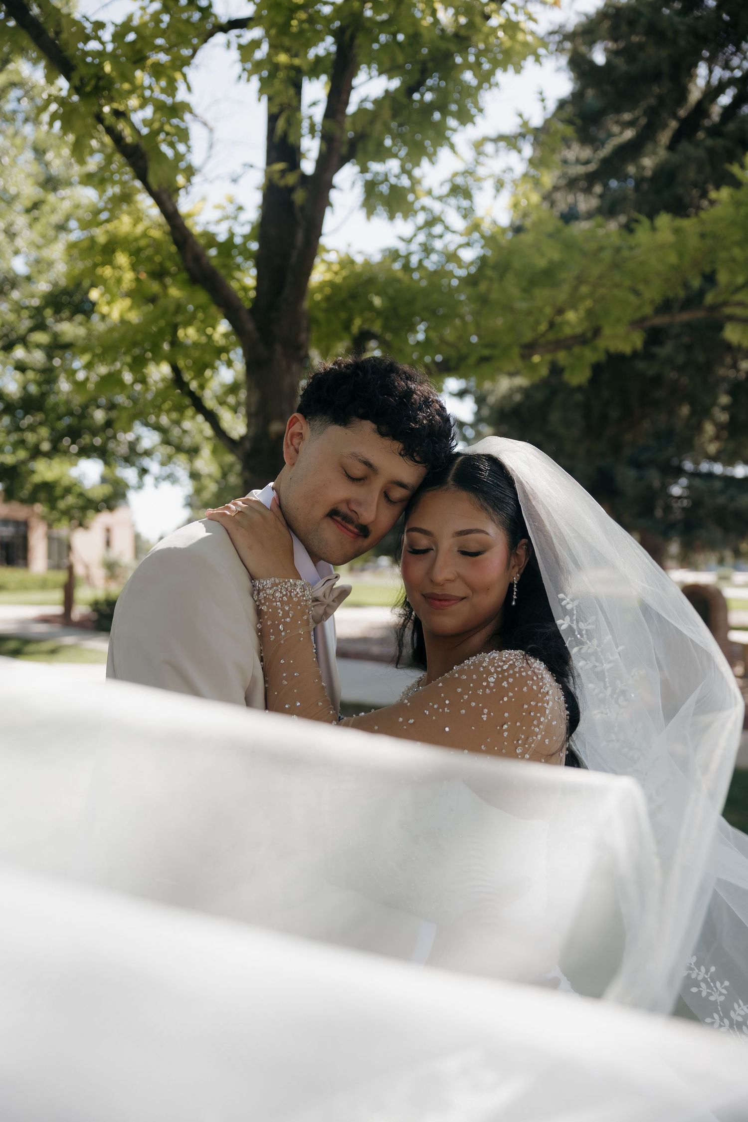 Bride and groom share a romantic embrace outdoors under trees in wedding attire.
