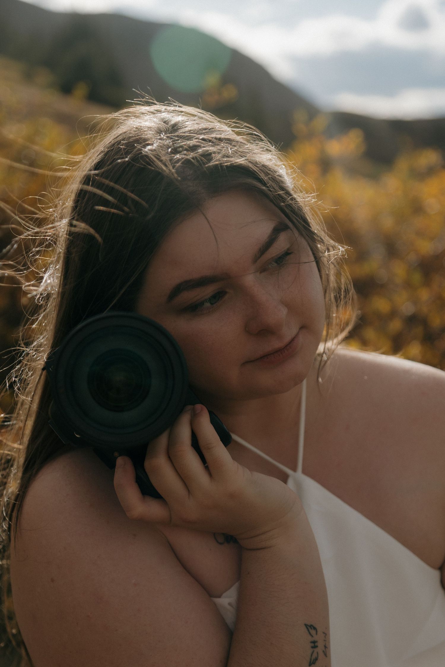 Young woman with long hair holds a camera lens to her cheek outdoors in a golden field with mountains behind her.