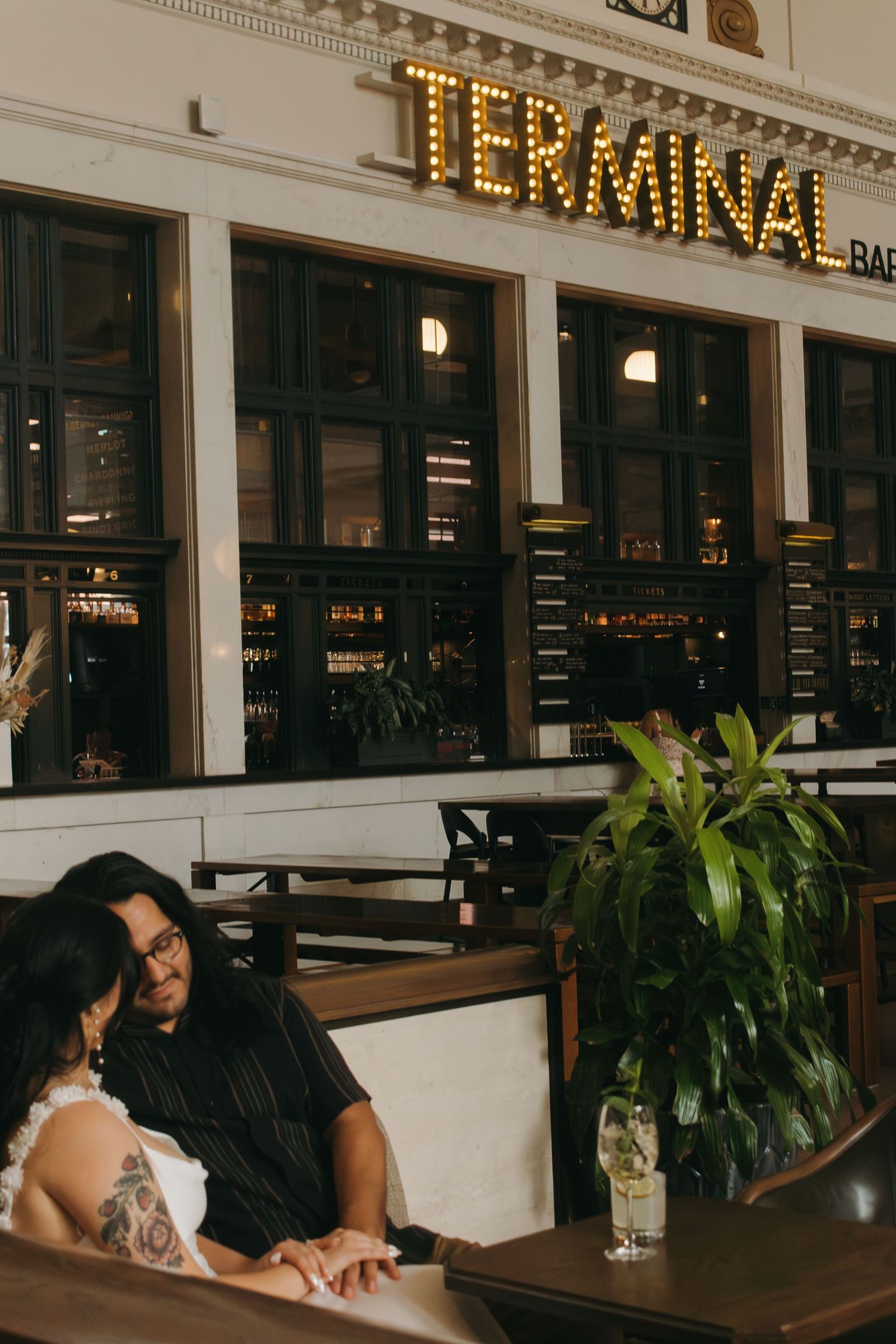 A couple shares an intimate moment at a restaurant booth with modern decor and plants in the background.