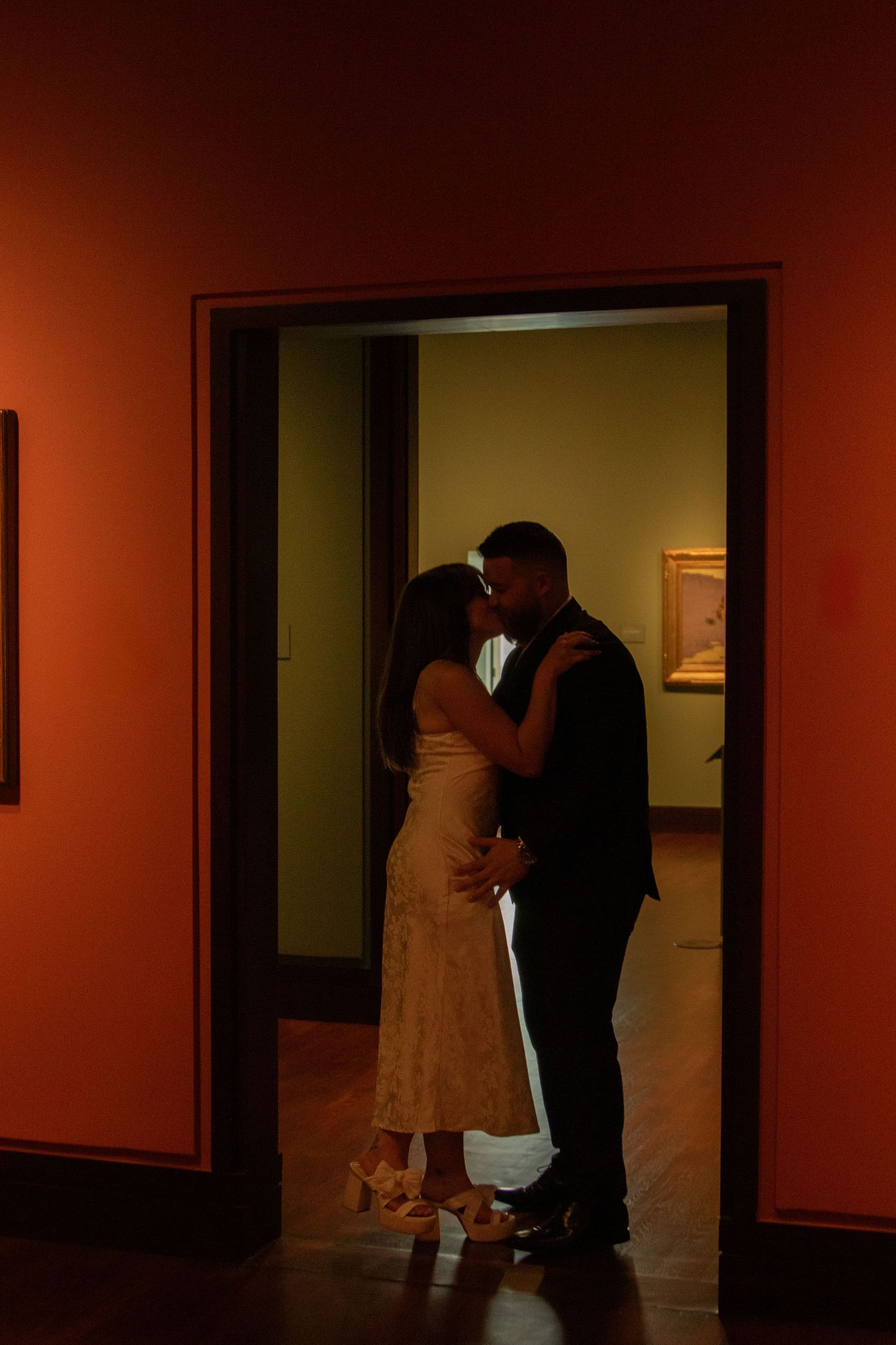 Two figures in dark hallway of art gallery with dim orange lighting.