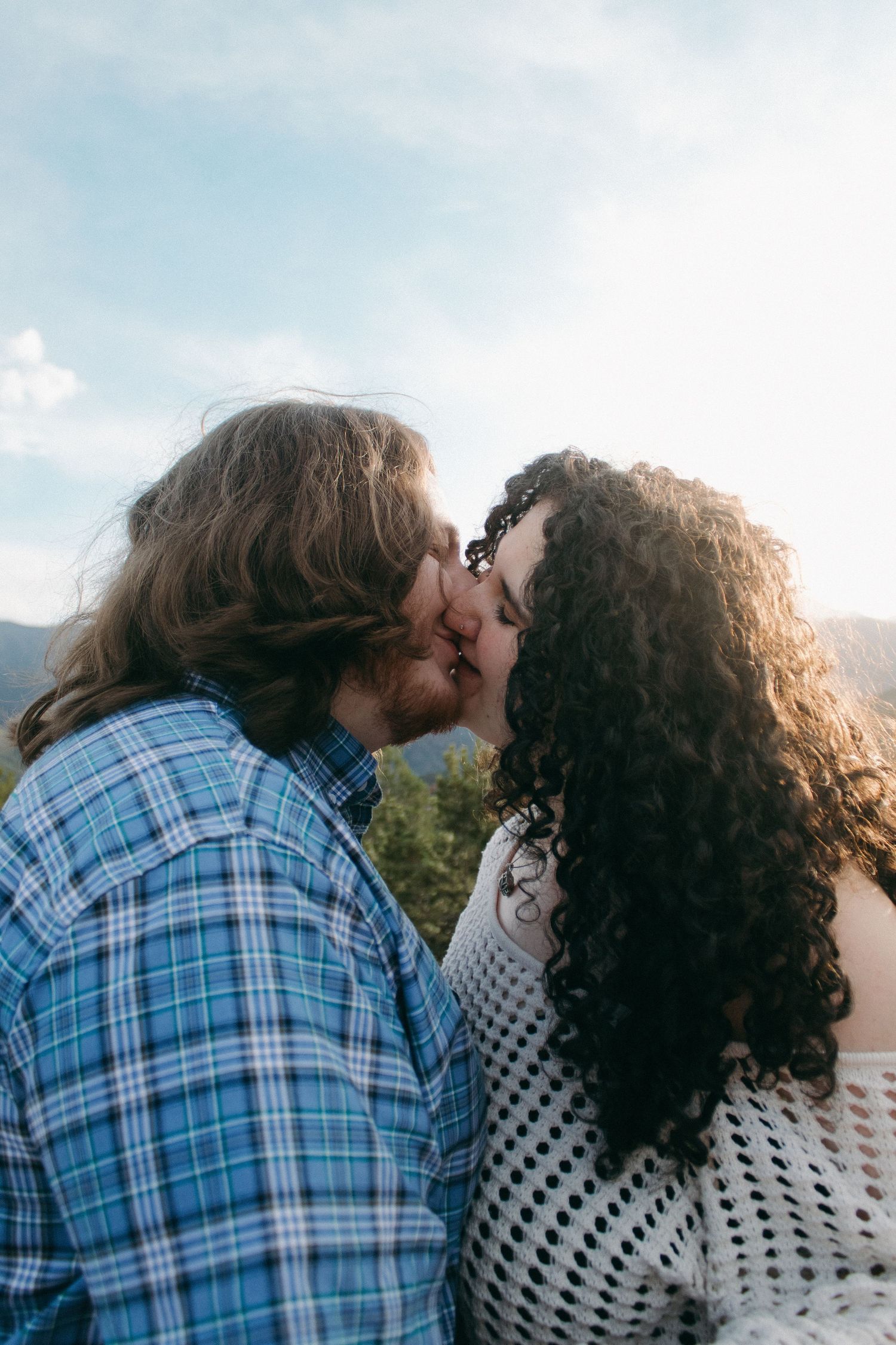 Two people in plaid and white sharing a tender kiss against a mountain landscape.