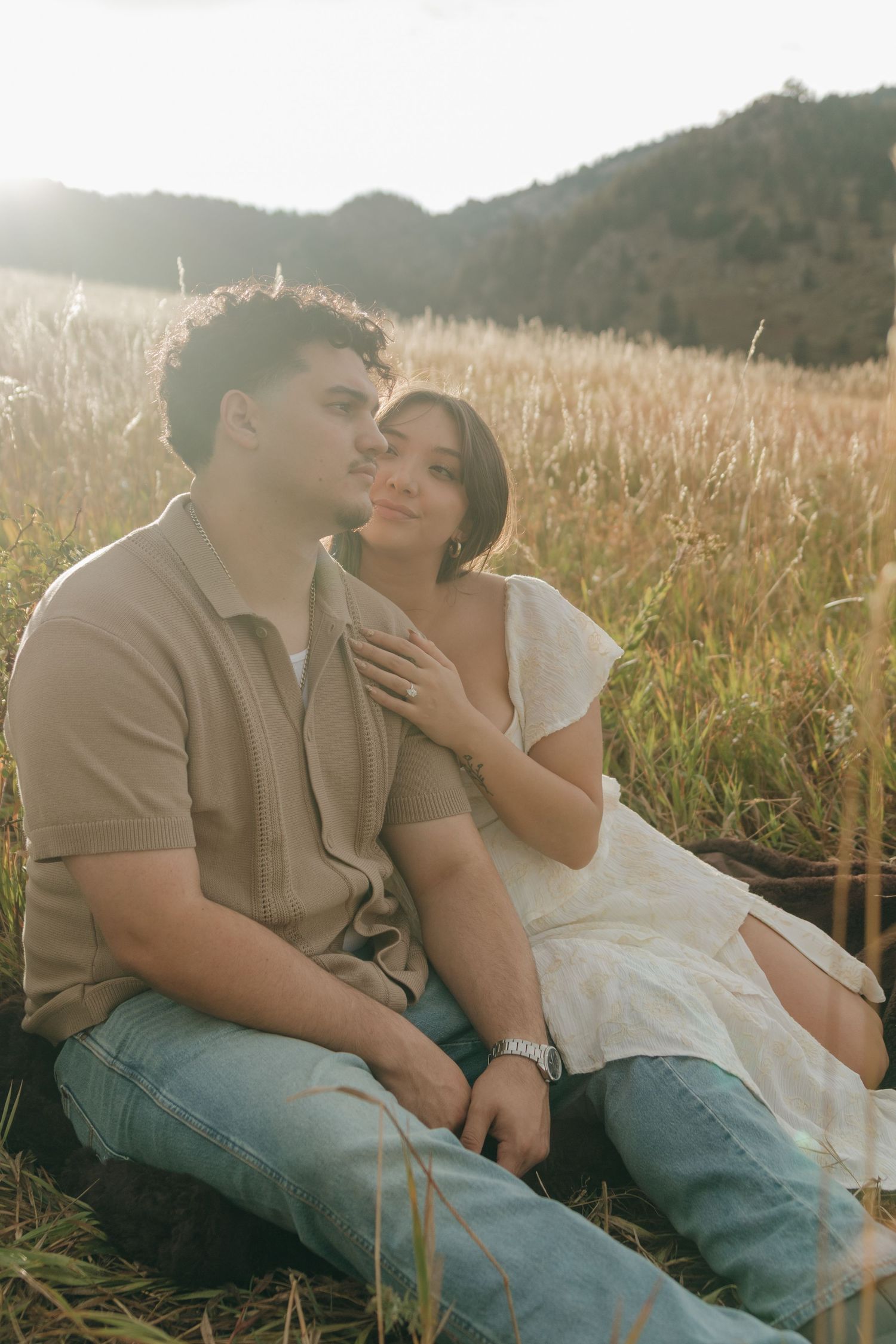 Couple sitting together in sunlit golden grass field with mountain views during engagement session.