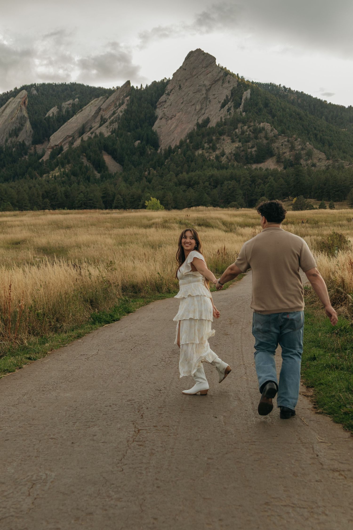 Couple holds hands walking on path toward dramatic Flatiron rock formations in Boulder.