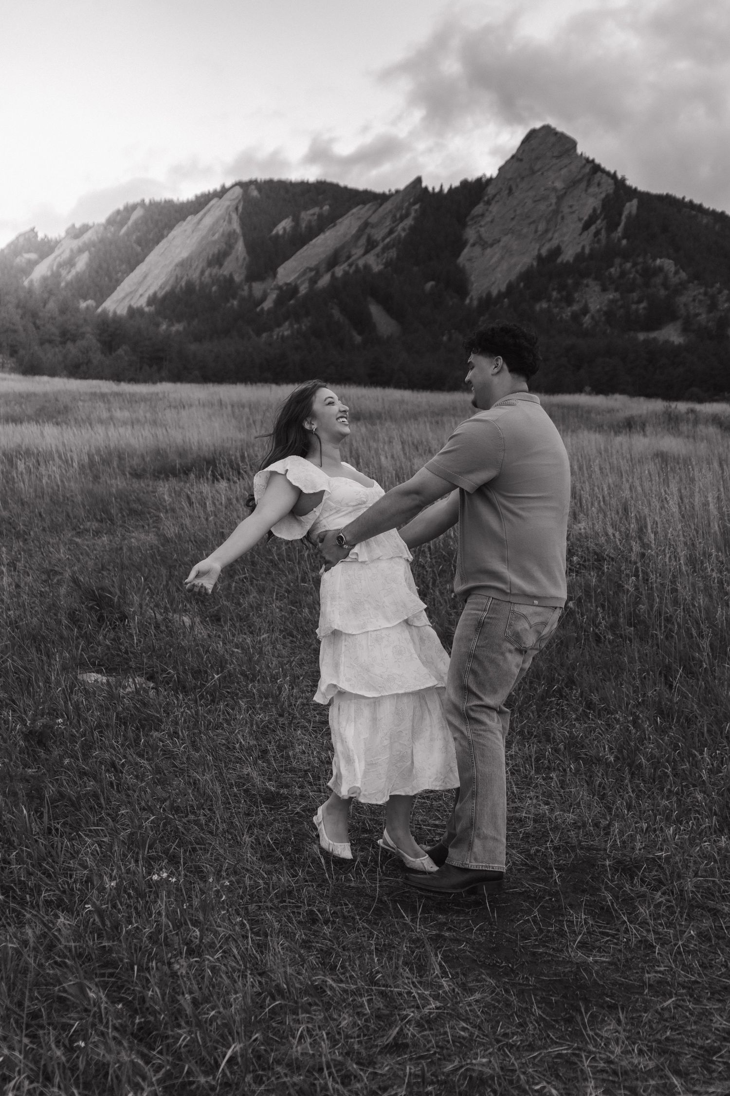 Black and white photo of joyful couple dancing and laughing together in field with mountain backdrop.