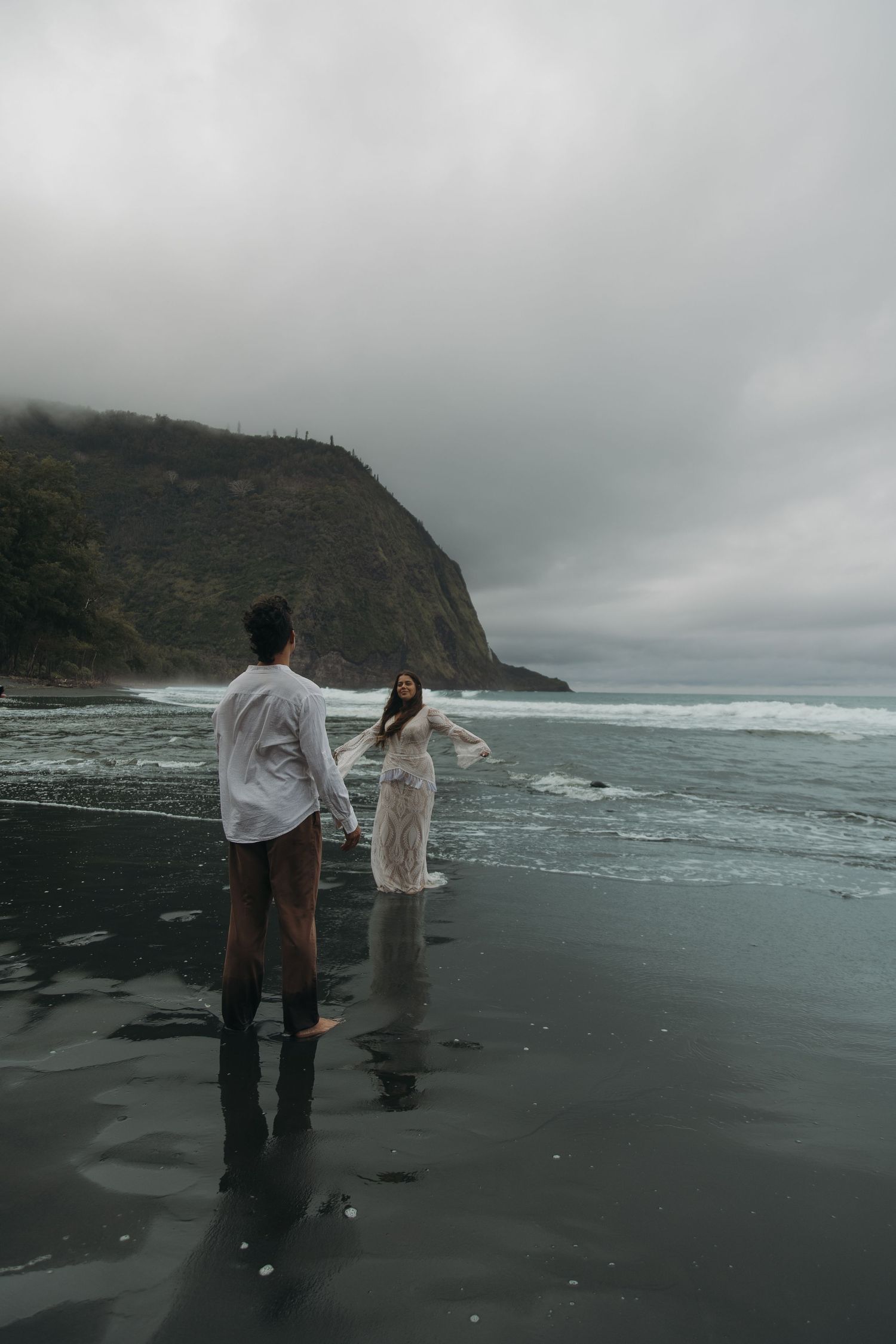 Couple holding hands on reflective black sand beach with misty cliffs and crashing waves in background.