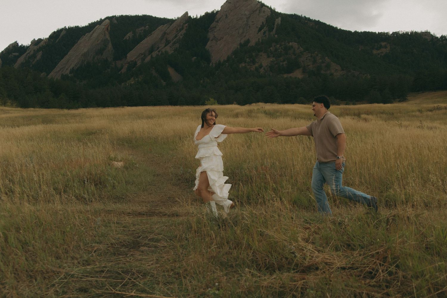 Couple holding hands running through golden field with Colorado Flatirons in background.
