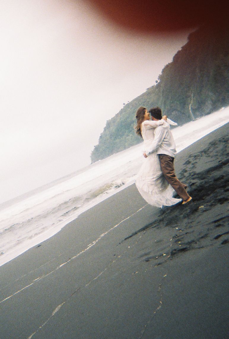 A couple shares a romantic embrace on a misty black sand beach, with ocean waves and a dramatic cliff in the background.