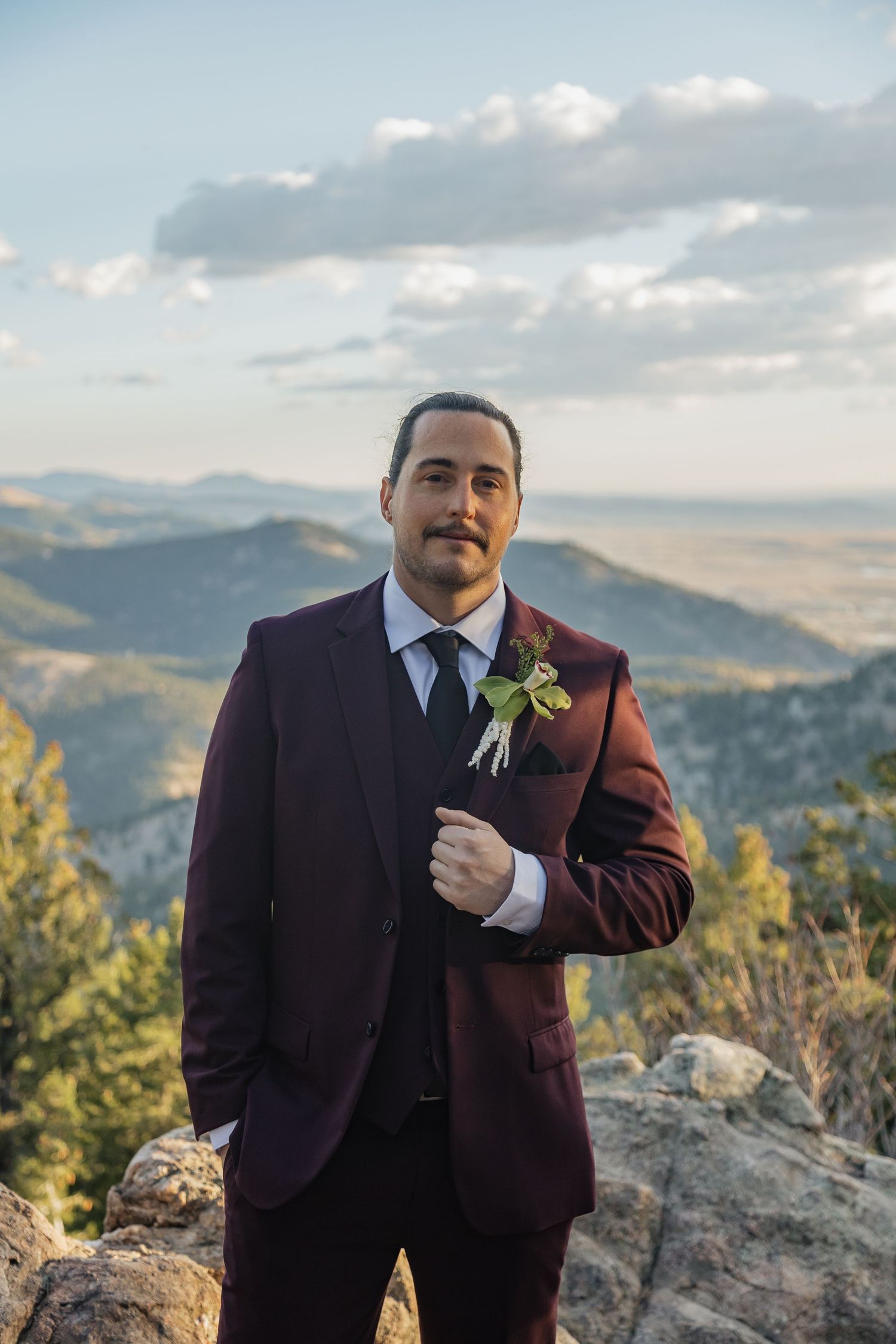 Groom in burgundy suit with boutonniere poses on rocky mountain overlook at sunset.