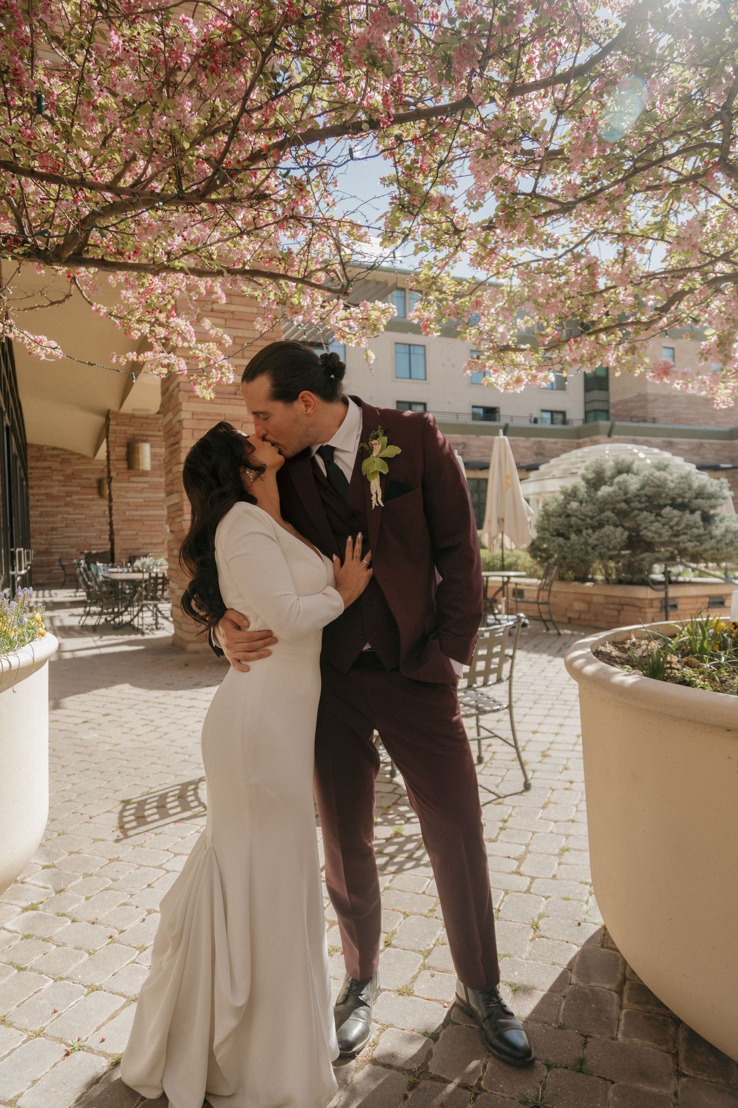 Bride and groom sharing romantic kiss under blooming pink cherry blossom tree on sunny wedding day.