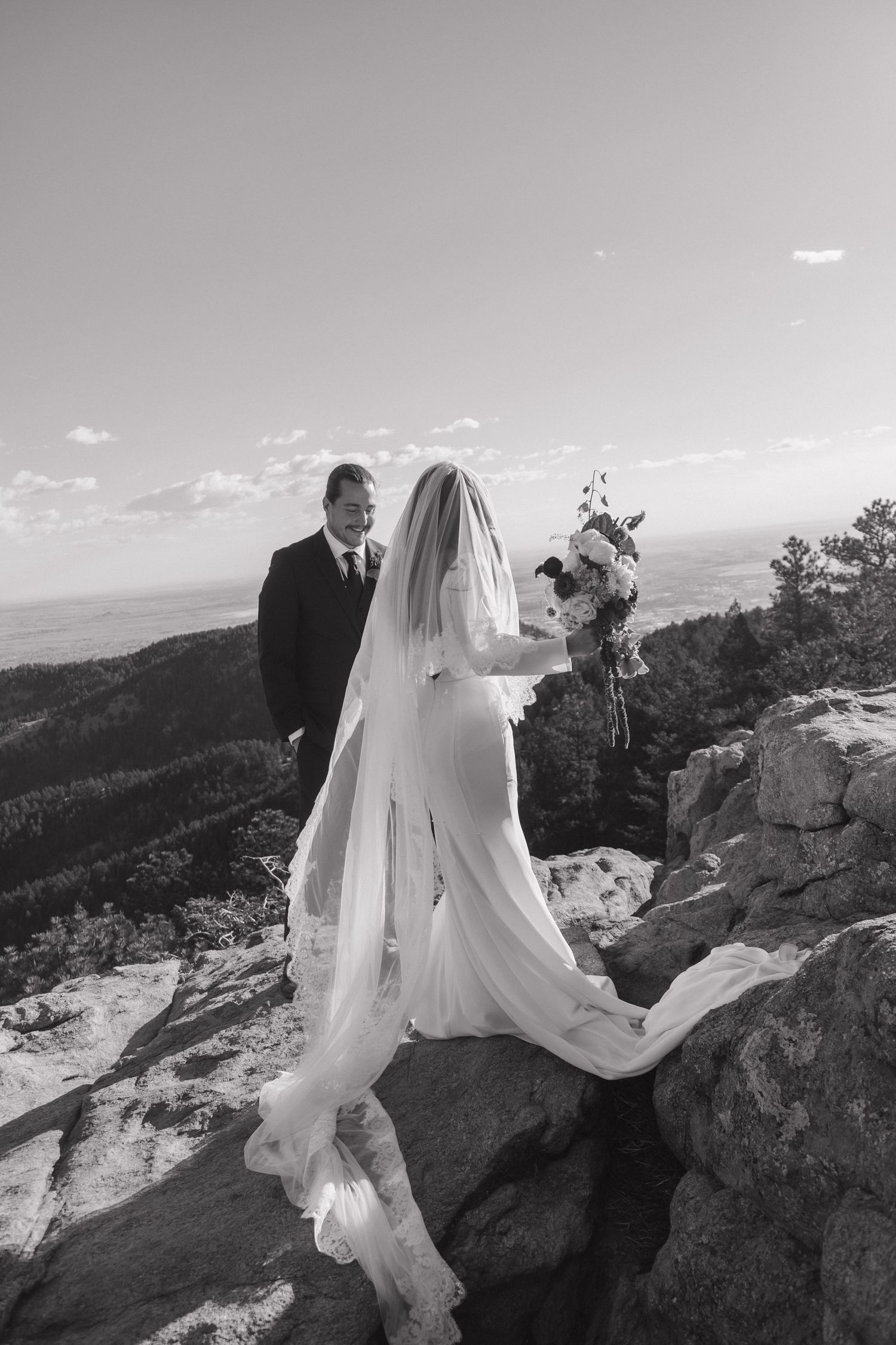 Black and white photo of bride in flowing veil and groom sharing moment on mountain cliff overlook.