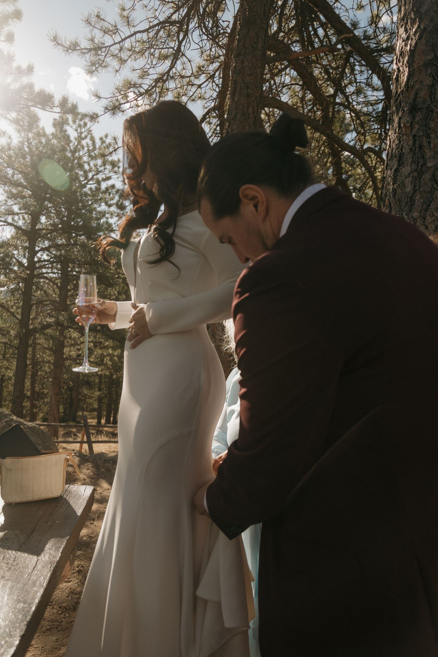 Bride in white gown holding champagne flute while groom in burgundy suit leans close in sunlit forest.