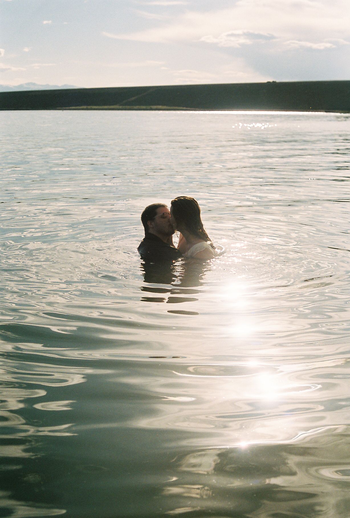 Couple kissing in open lake water with sun glare reflecting on the surface.