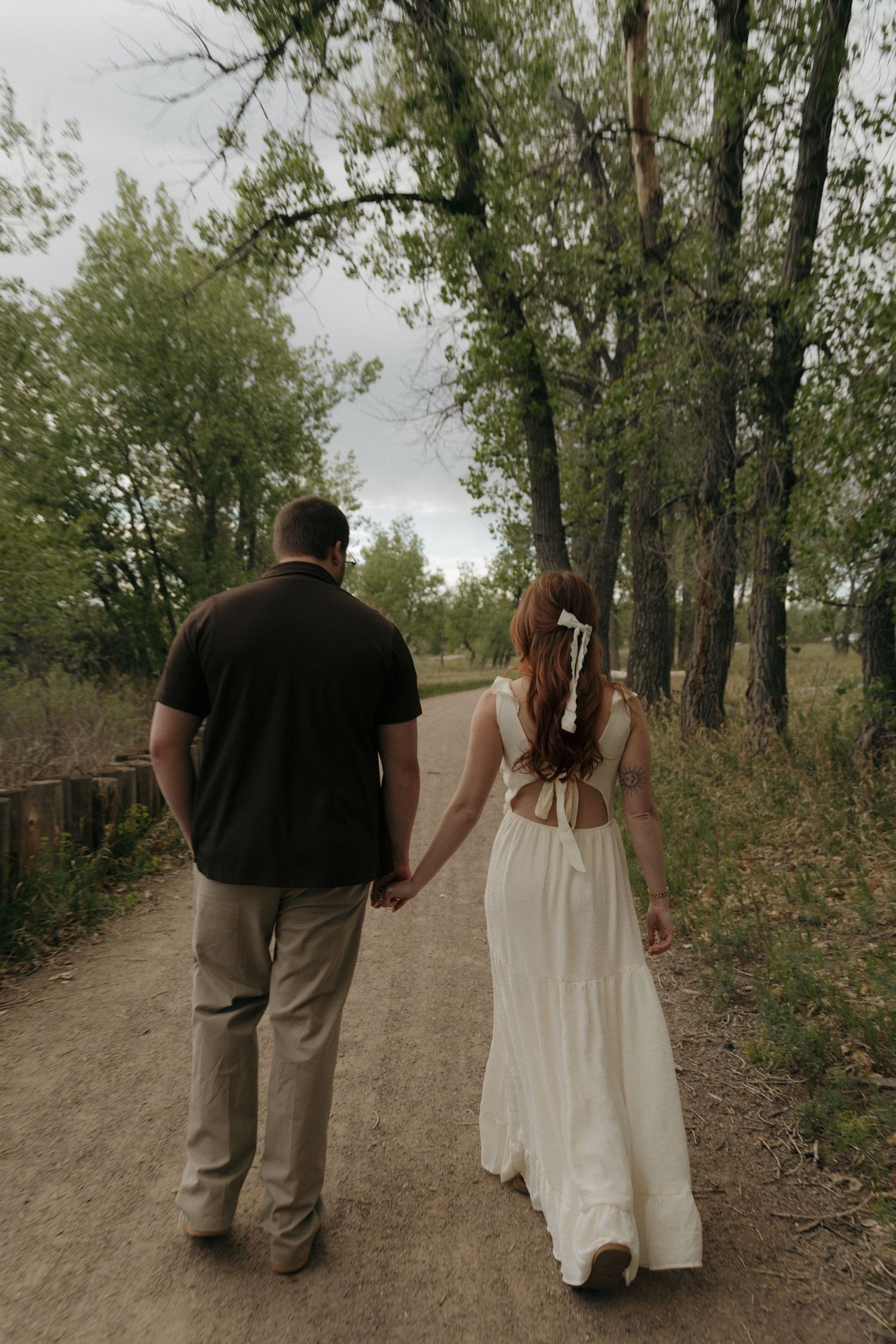 Couple holding hands walking down a tree-lined dirt path, woman in white dress with bow hair tie.