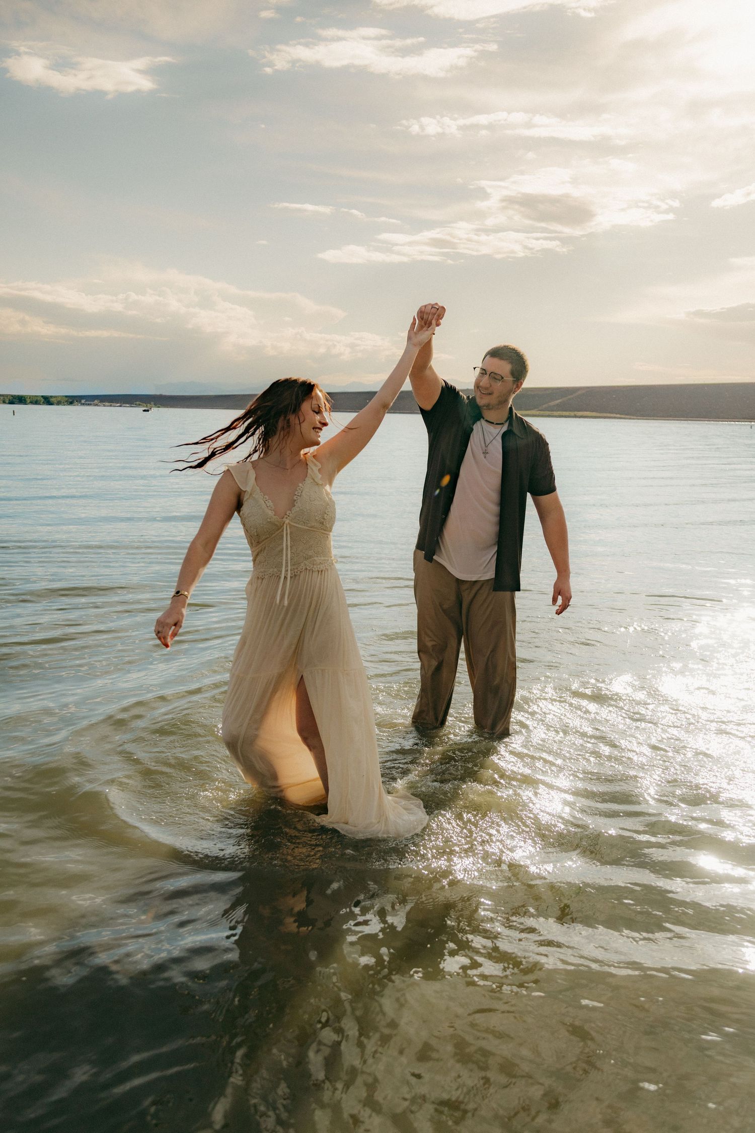 Joyful couple holding hands and walking through shallow lake water at golden hour sunset.
