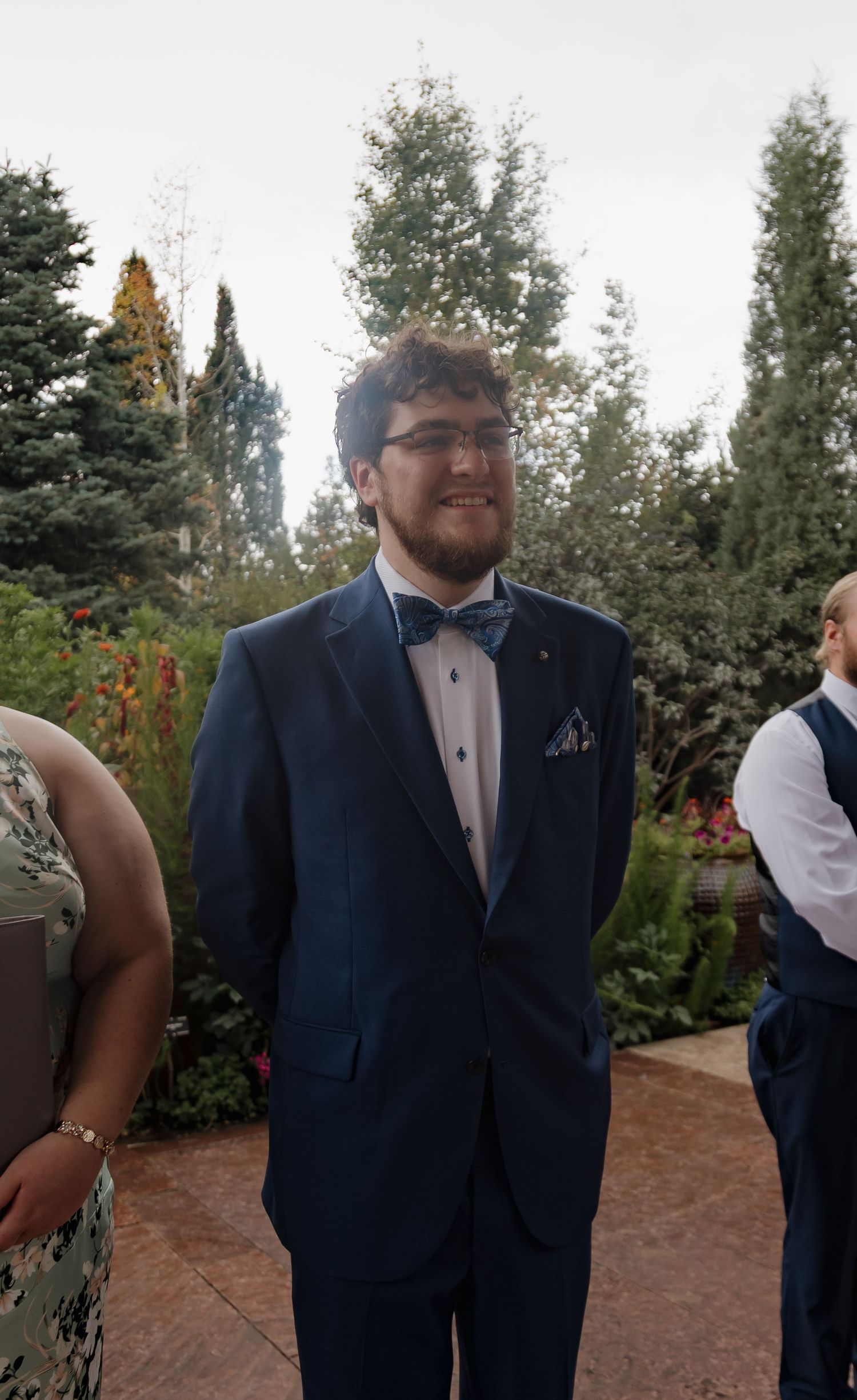 Smiling groom in navy blue tuxedo with bow tie and pocket square standing outdoors at a garden wedding.