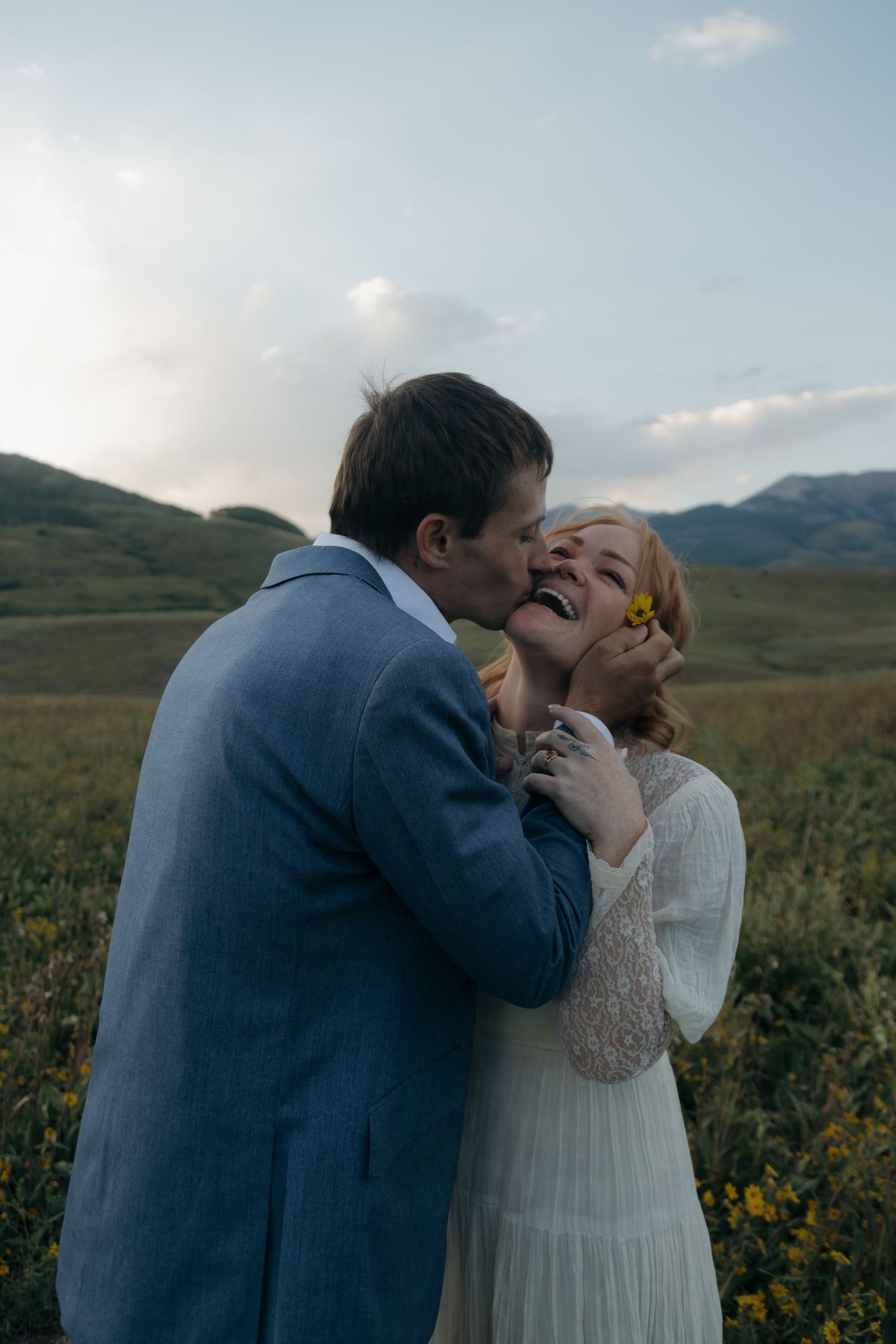 Groom in blue suit kisses laughing bride in white dress in wildflower meadow with mountain backdrop.