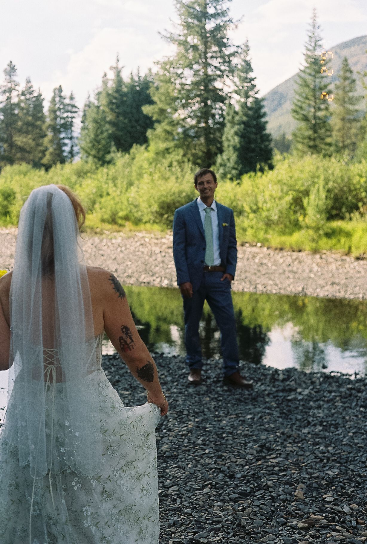 Bride with arm tattoo in lace gown approaches groom in blue suit during first look by mountain creek.