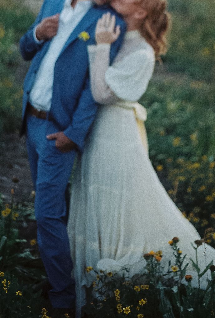 A couple embraces in a wildflower field, bride in white gown with yellow sash, groom in blue suit.