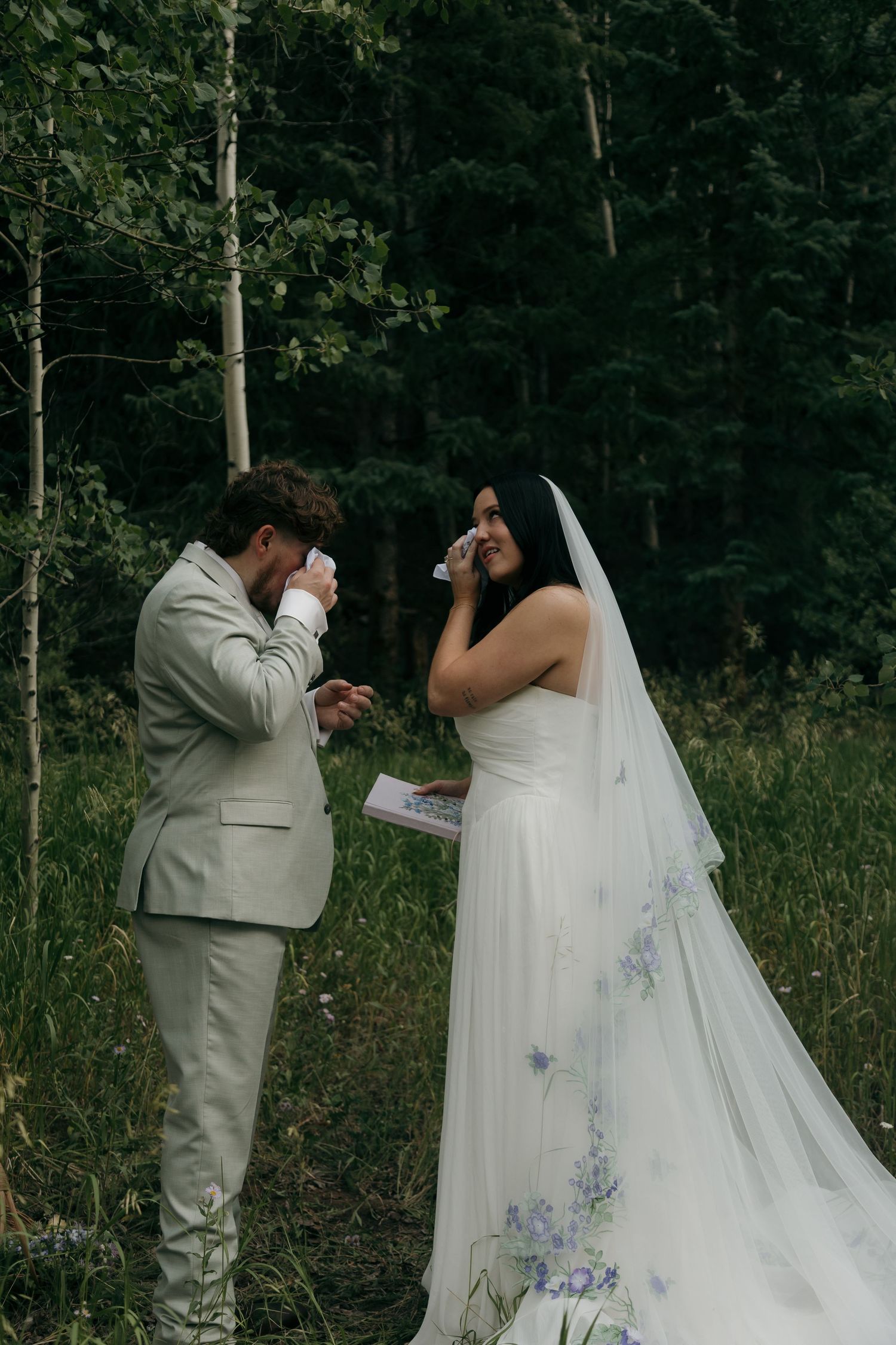 Bride and groom wiping tears during emotional outdoor forest wedding vow exchange.