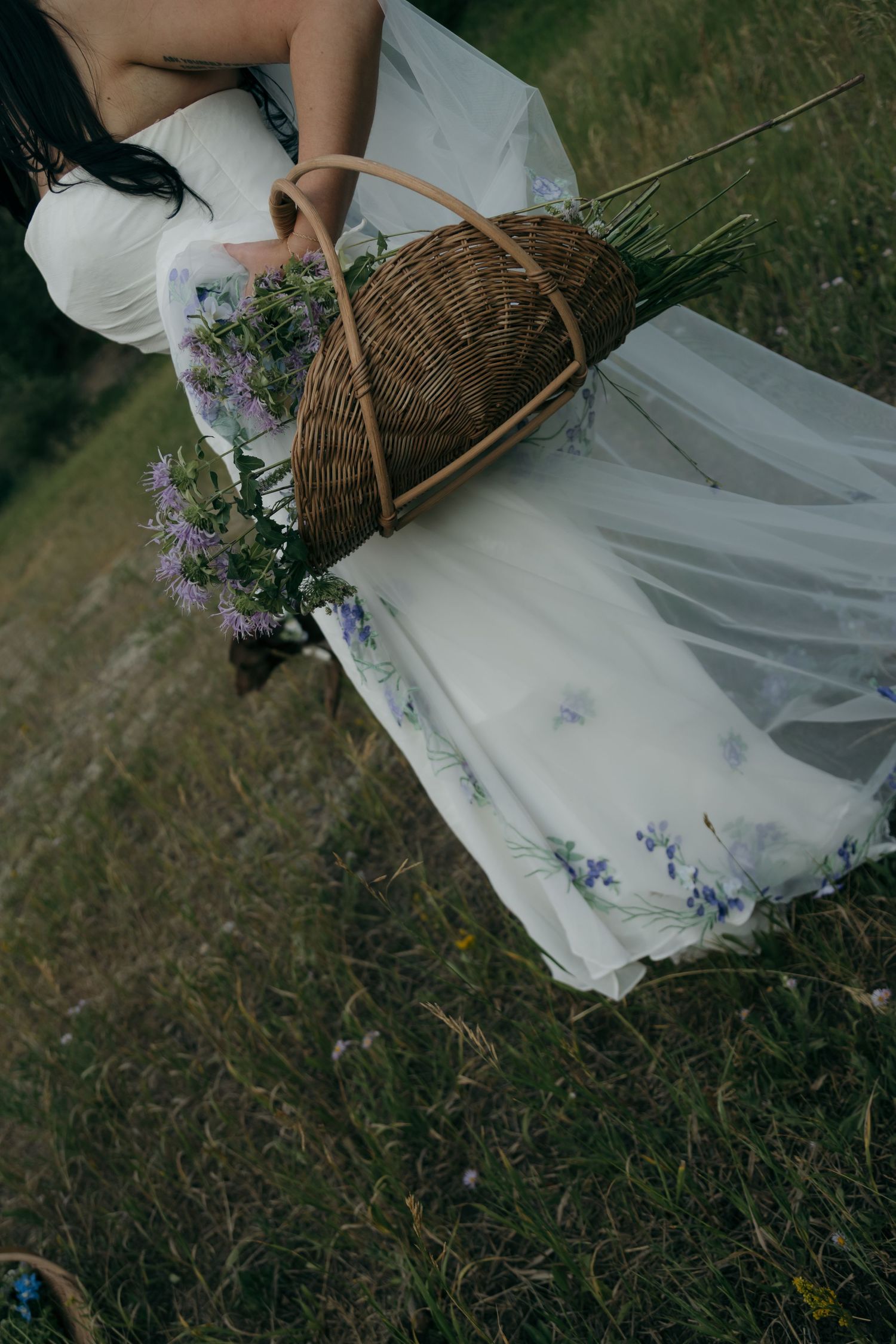 Bride in white floral wedding gown carries rustic wicker basket with wildflowers in meadow.
