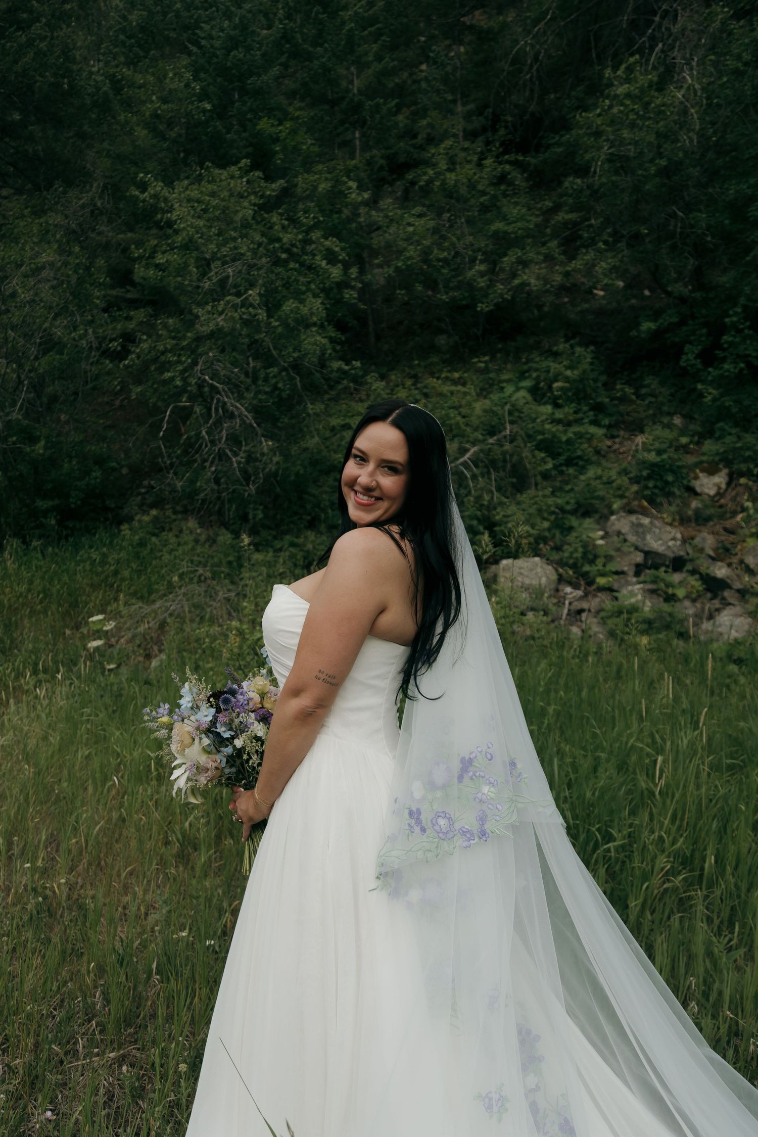 Bride in white gown with floral veil smiles over shoulder holding wildflower bouquet.