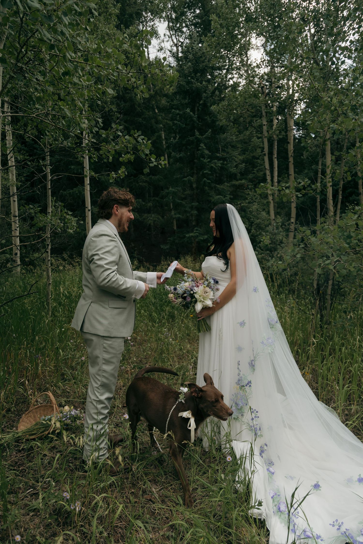 Groom reading vows to bride with floral veil beside dog in forest wedding ceremony.