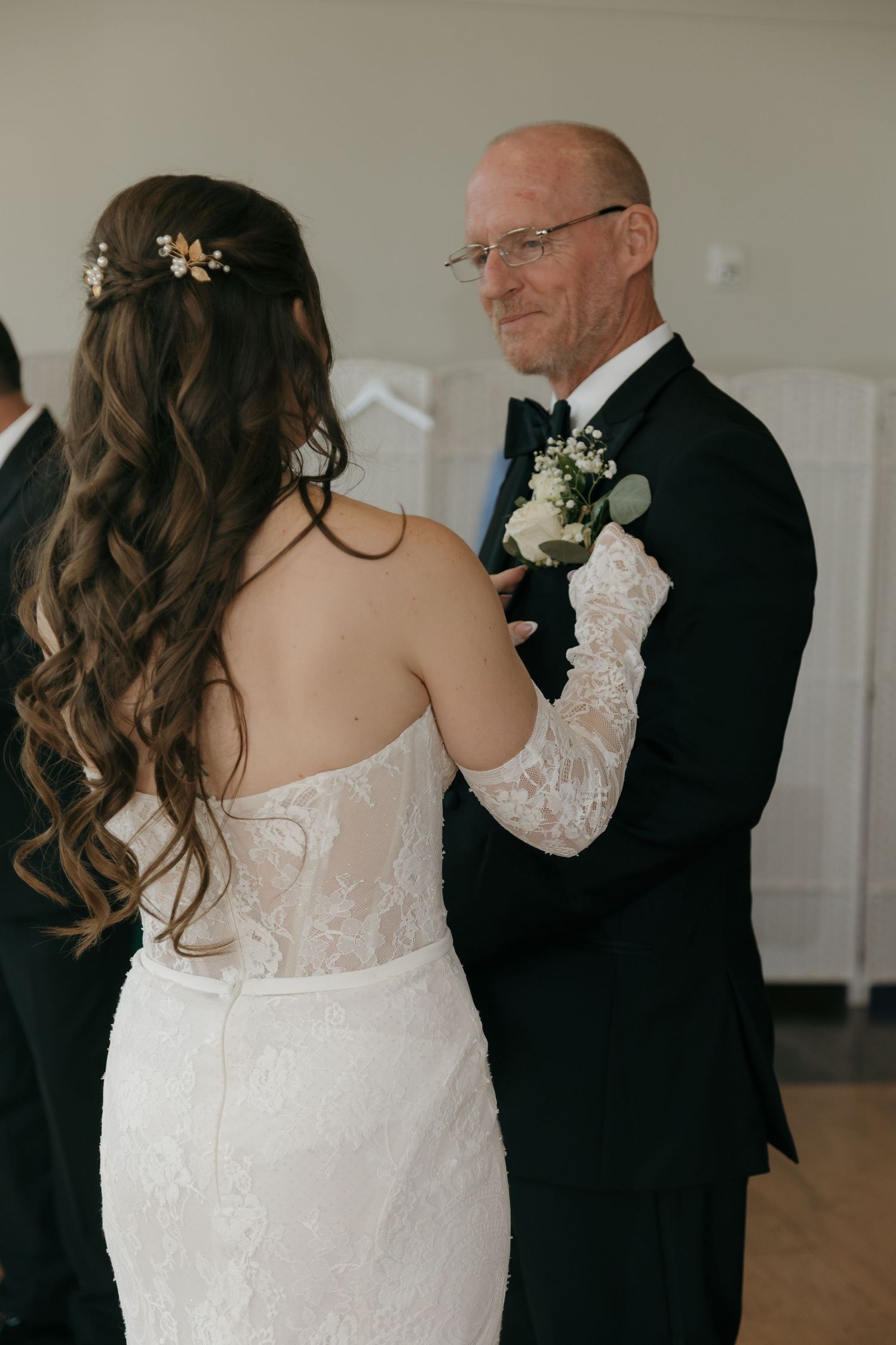 Bride in lace gown pinning boutonniere on father in tuxedo before wedding ceremony.