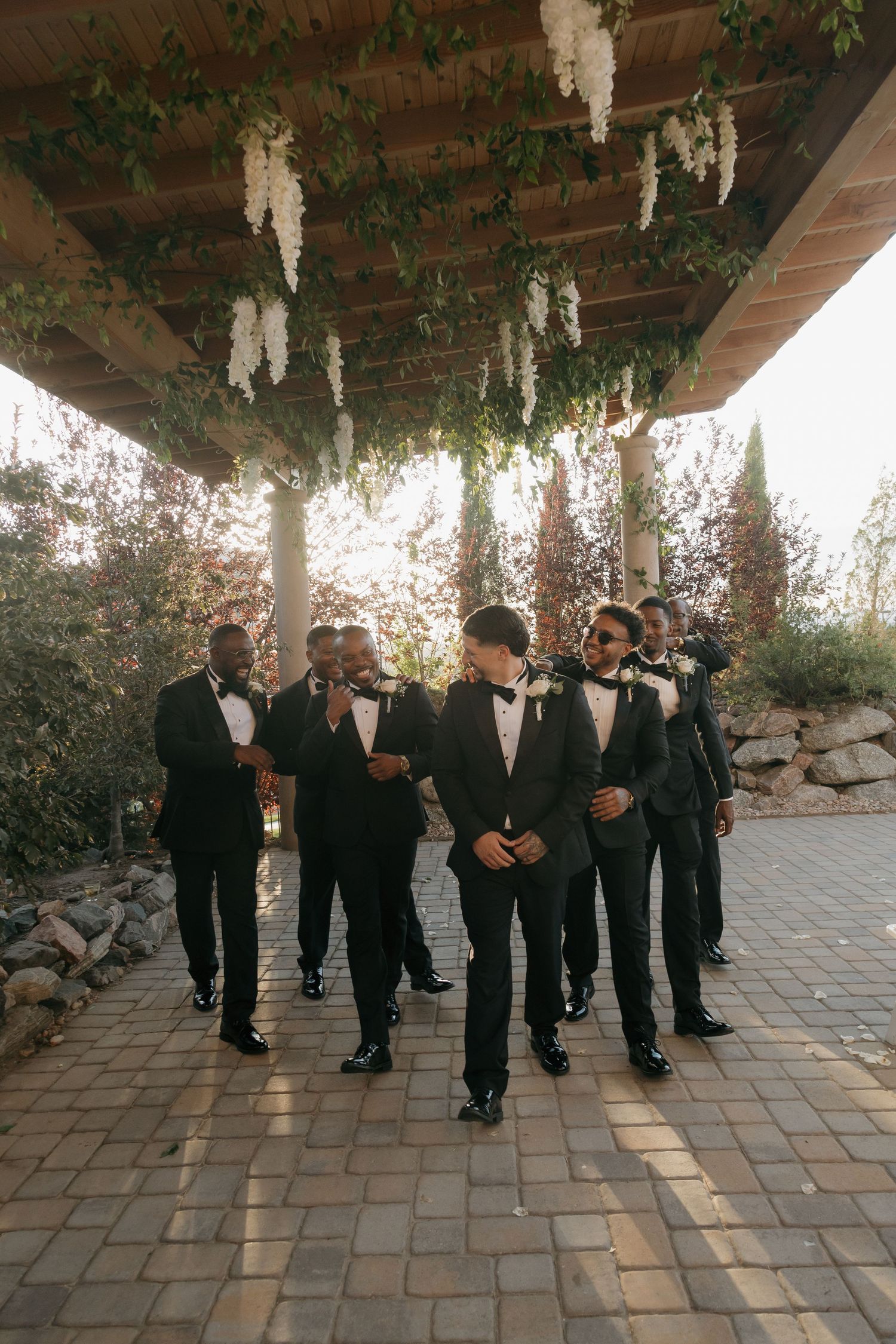 Groomsmen in black tuxedos walk together under a floral pergola with hanging white wisteria at a wedding.
