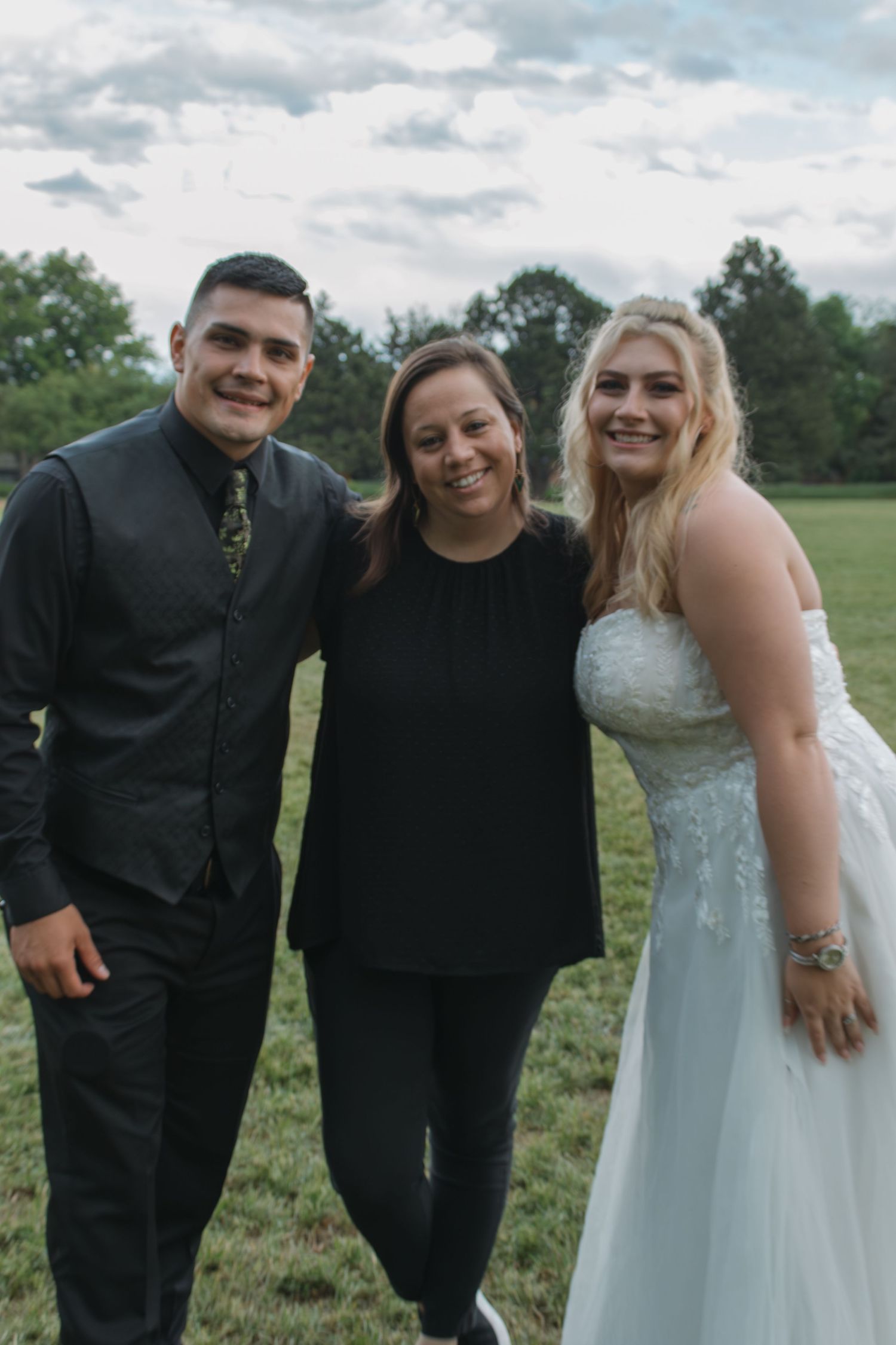 Wedding photo of three people posing together outdoors on grassy field.