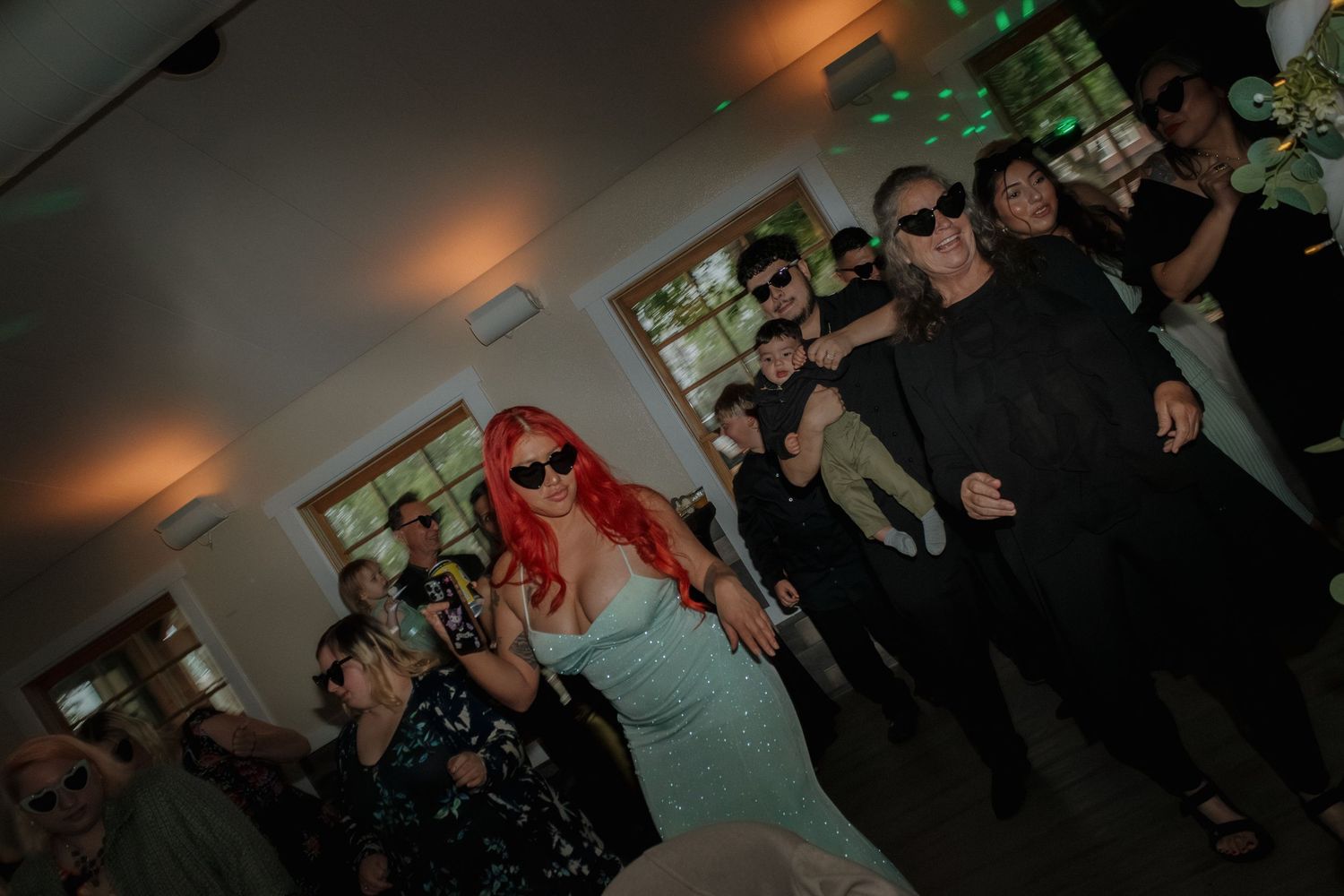 Crowded dance floor at a party with colorful lighting effects and windows in background.
