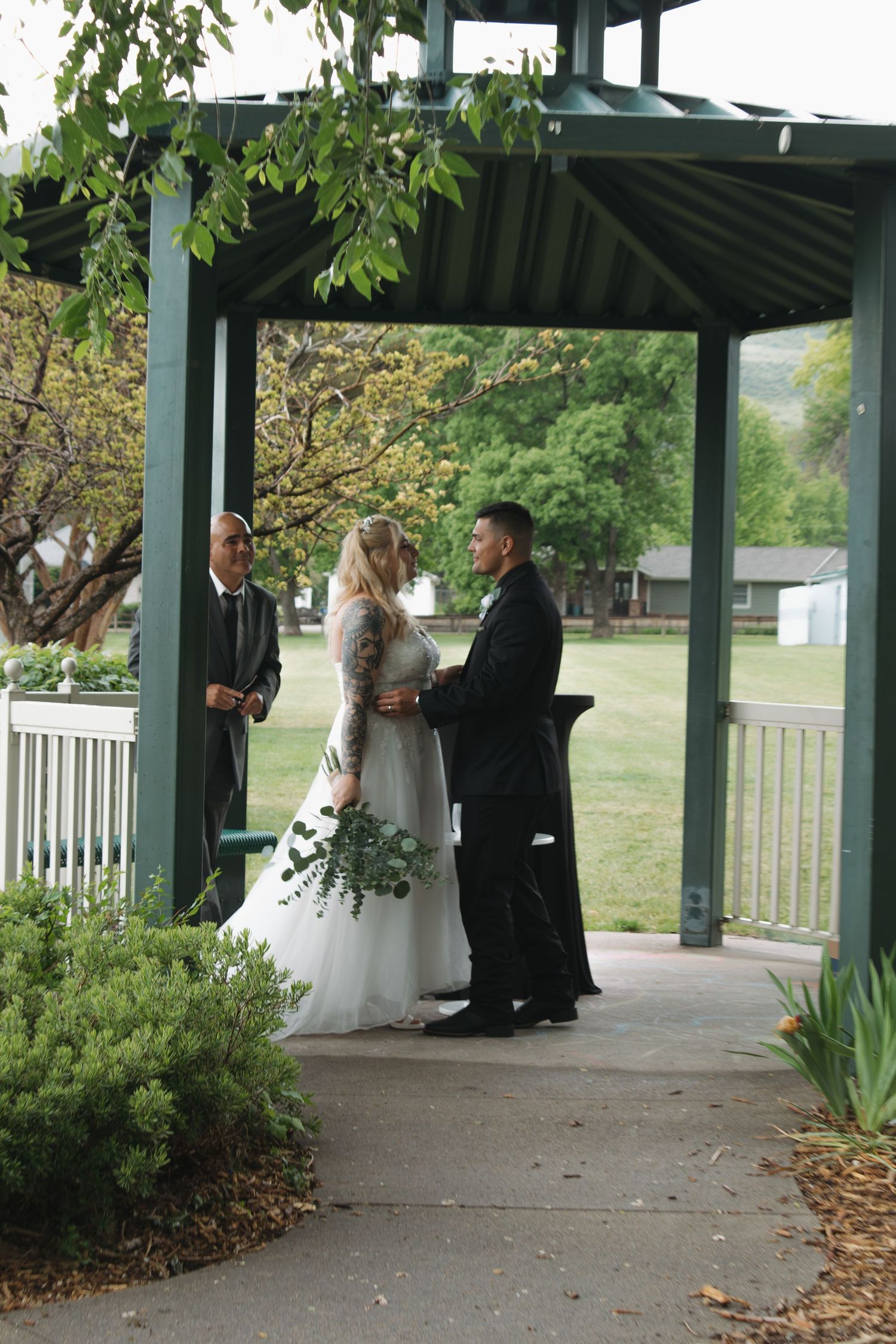 An outdoor wedding ceremony unfolds beneath a covered green pavilion surrounded by trees.