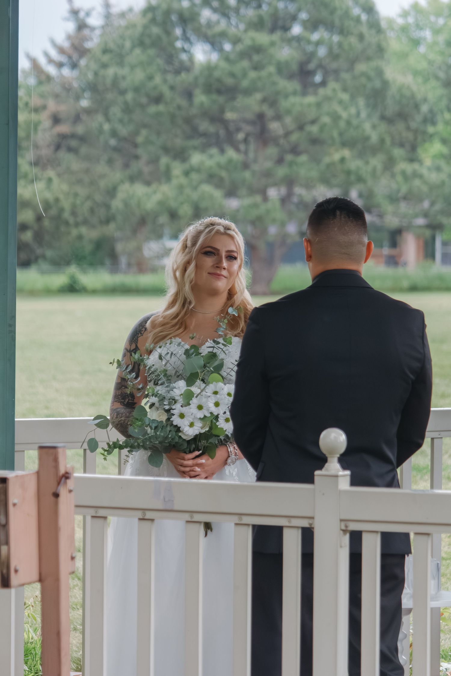 A couple stands together during their wedding ceremony on a covered porch surrounded by greenery.