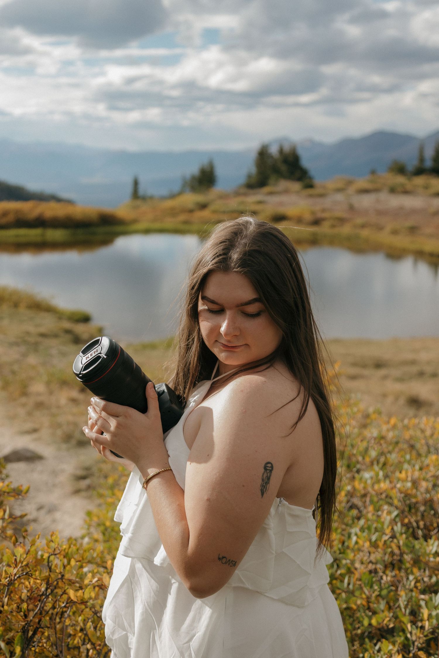 Young woman in white dress holds camera near mountain lake with cloudy sky and autumn foliage backdrop.