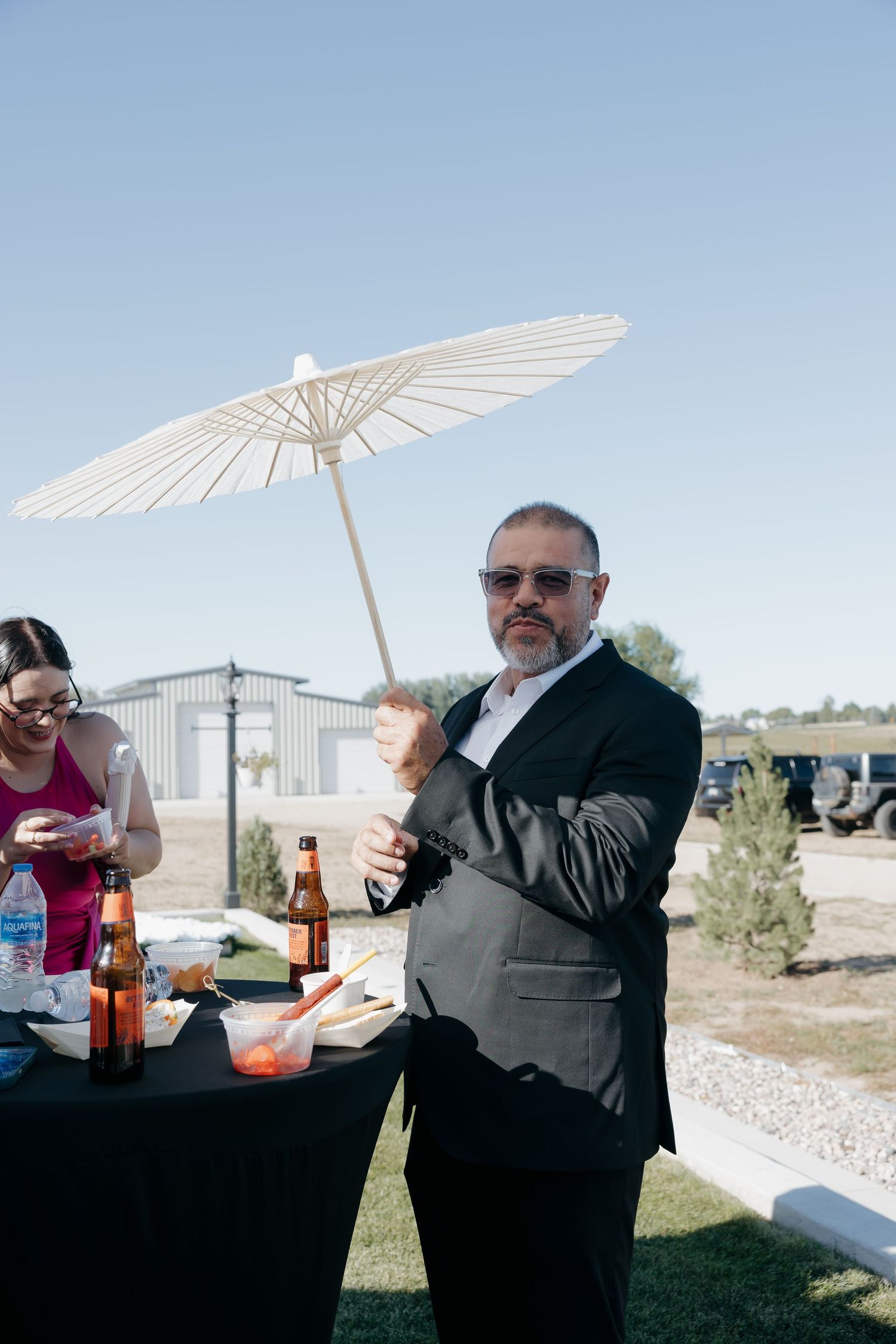 Man in dark suit holds white paper parasol at outdoor wedding reception table.