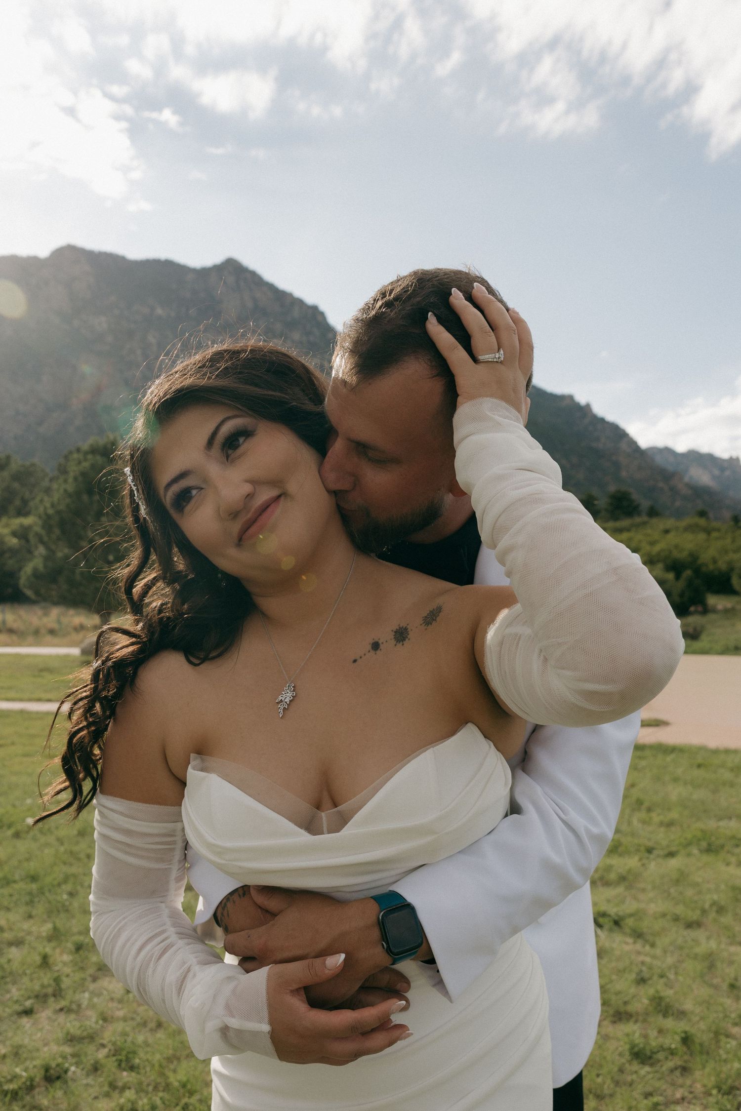 Groom embraces bride from behind in front of a scenic mountain landscape on wedding day.