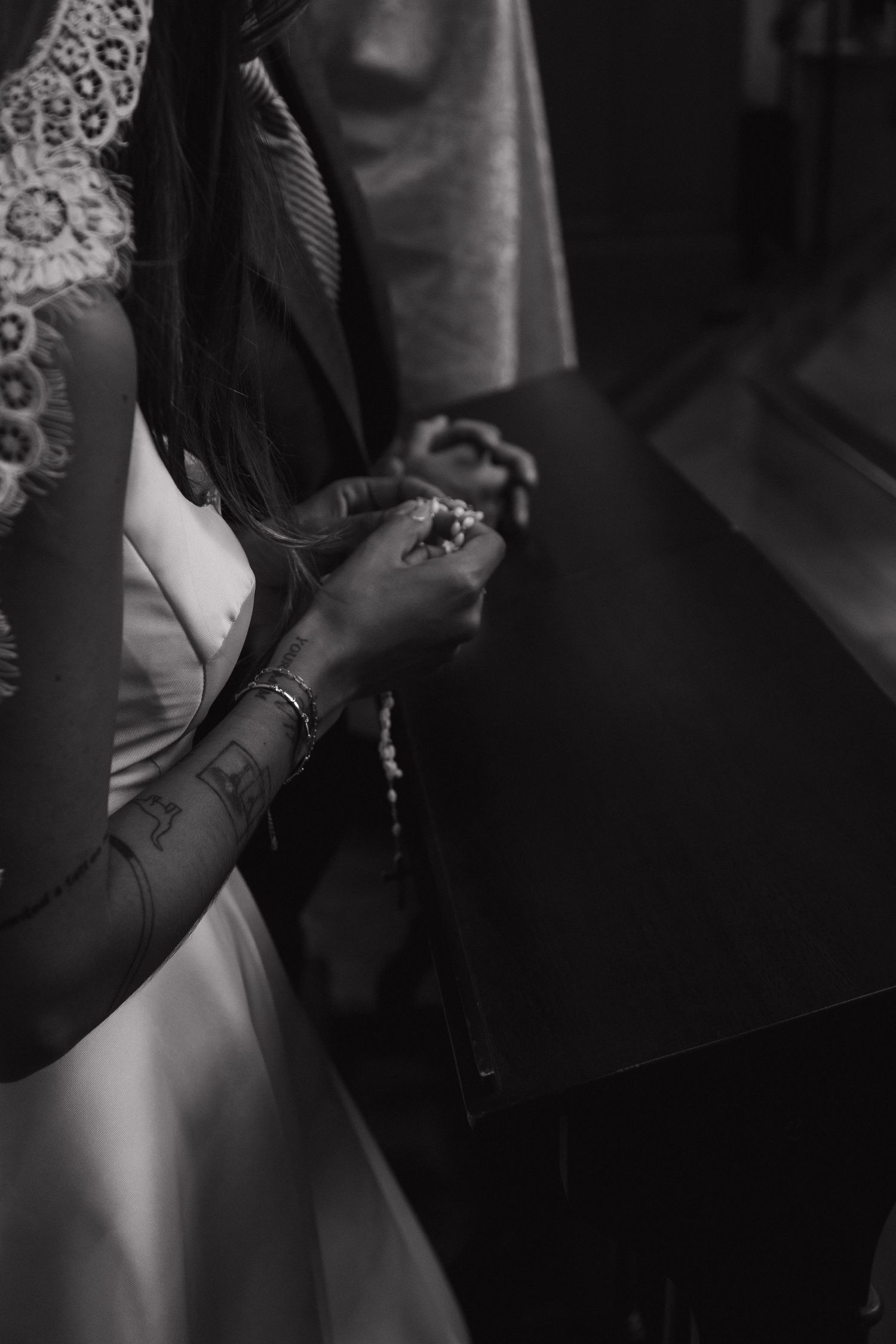 A bride in a lace wedding gown holds rosary beads while kneeling in prayer at a pew.