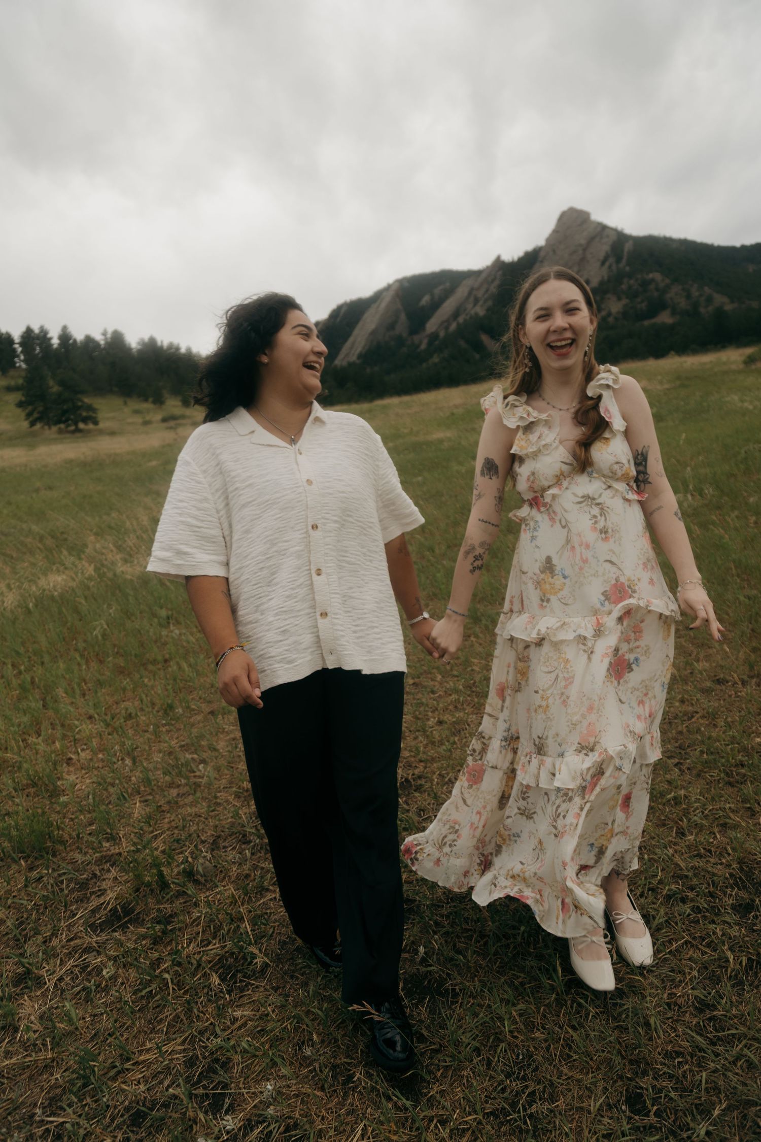 Happy couple holding hands and laughing while walking through a green meadow with rocky Flatirons backdrop.