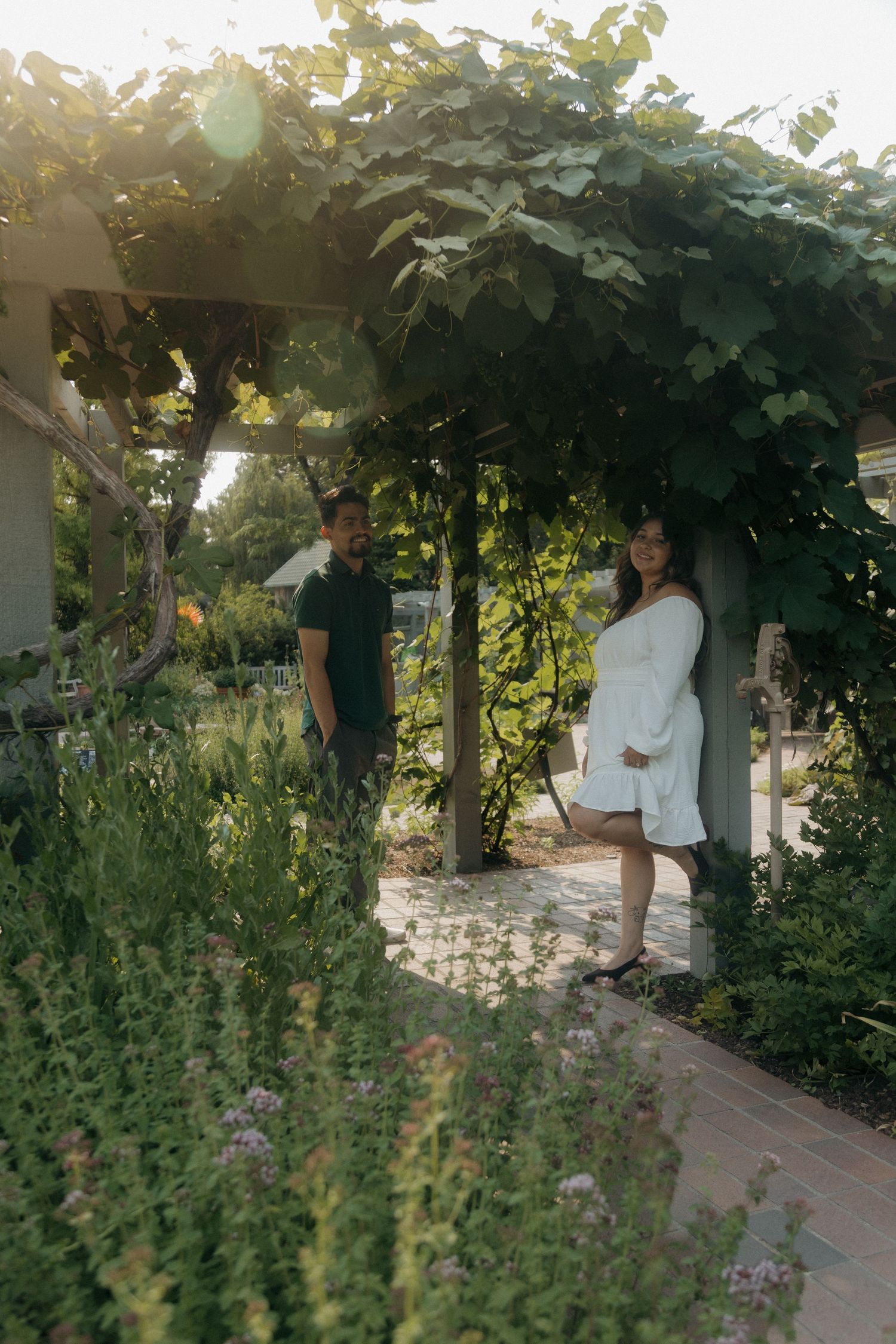 Couple standing under a vine-covered pergola in a sunlit garden, woman in white dress smiling at camera.