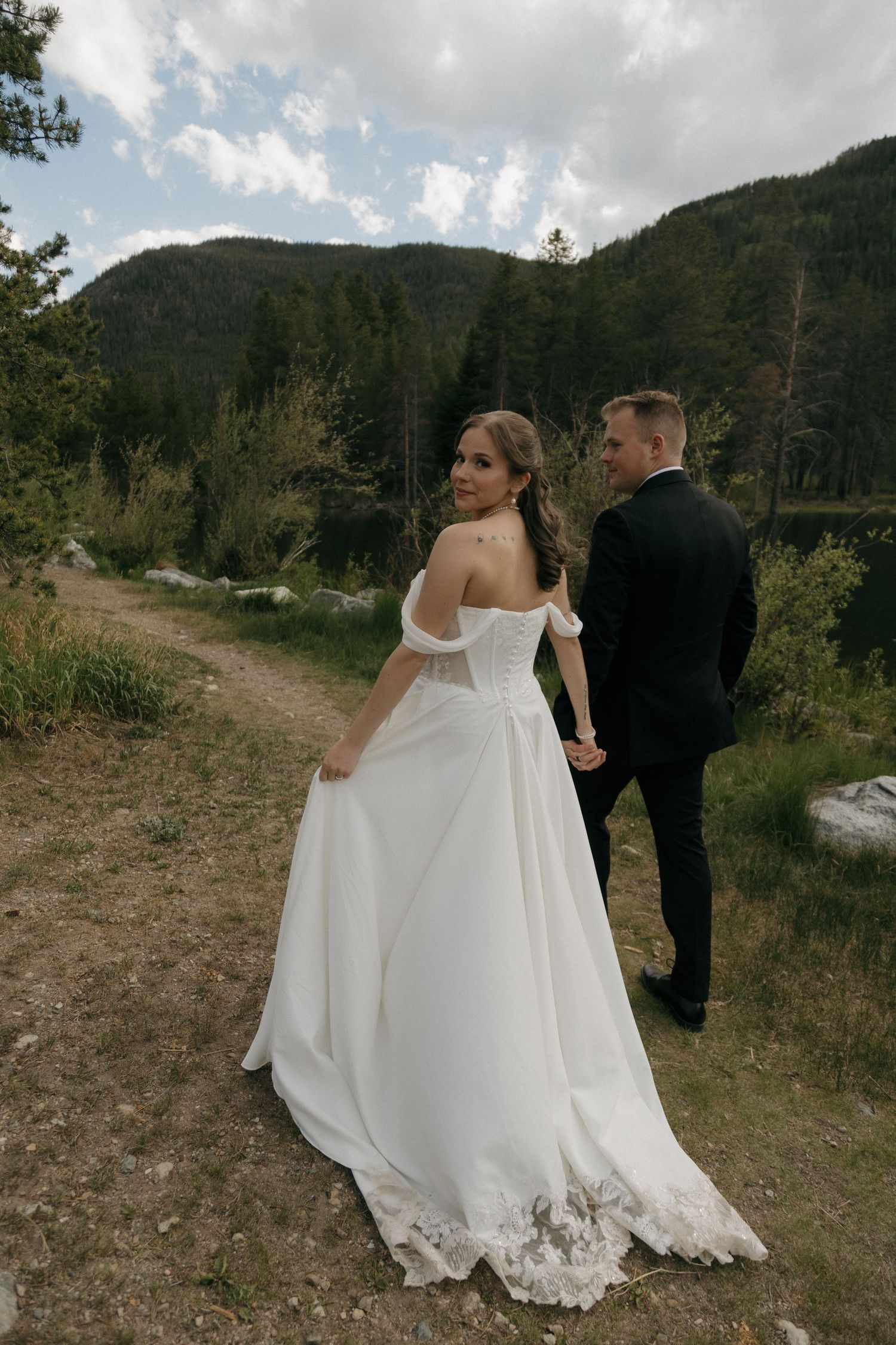Bride in off-shoulder white gown glances back while holding groom's hand on a mountain trail.