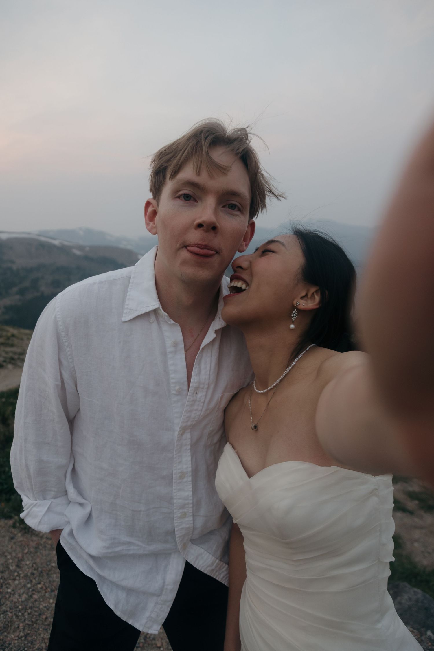 Joyful couple takes a selfie at golden hour on a mountain overlook, bride in strapless gown laughing warmly.