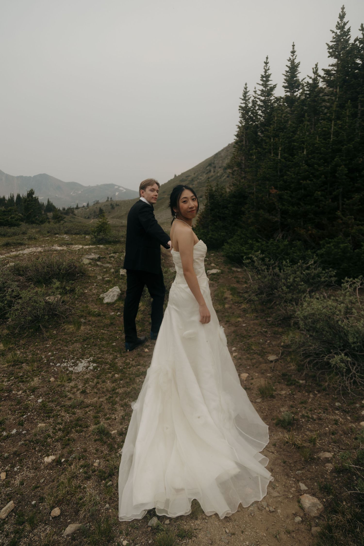 Bride in flowing white gown looks back smiling while groom in tuxedo holds her hand on a mountain trail.