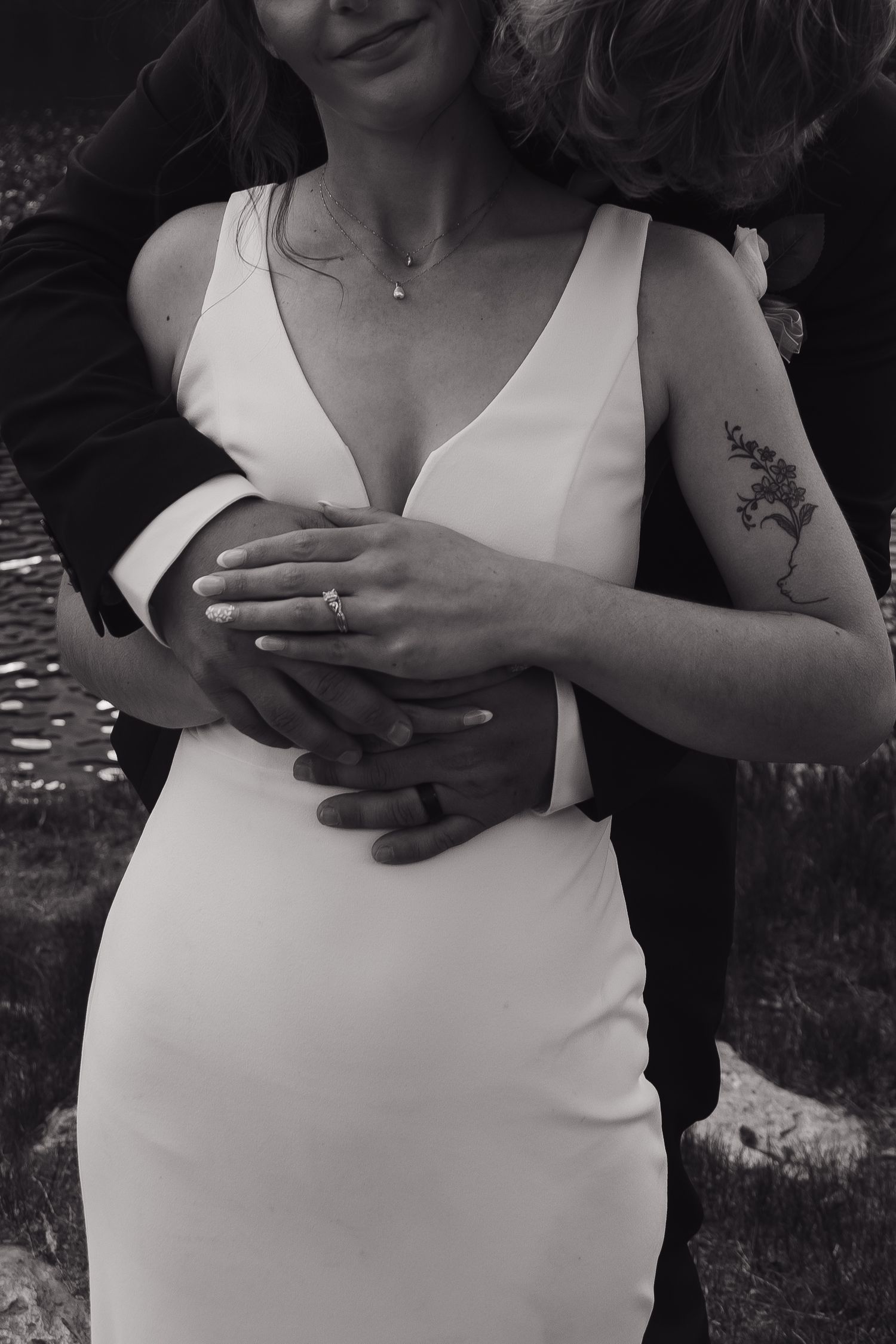 Black and white photo of a bride in a white gown embraced by groom, showing wedding ring and floral tattoo.