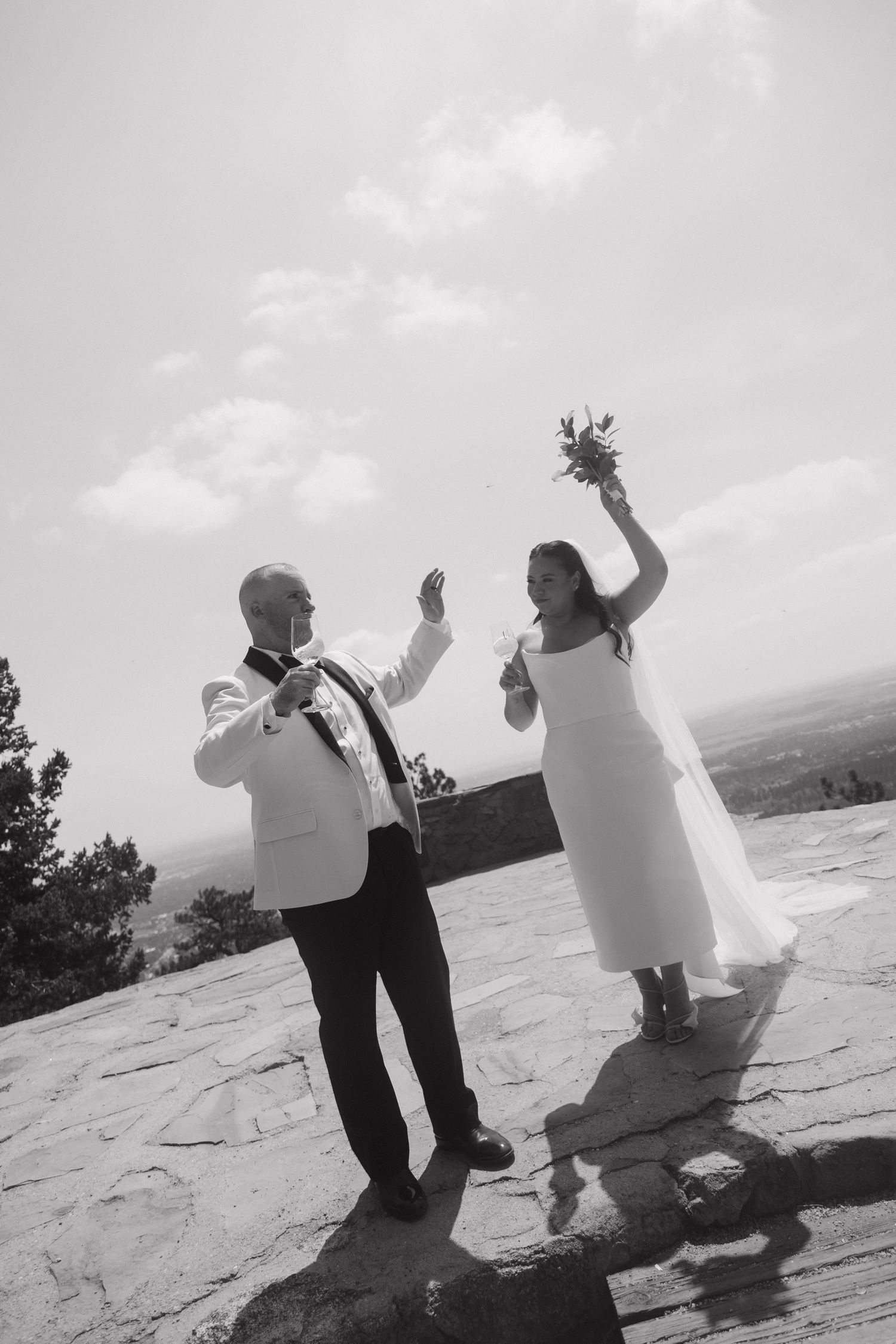 Black and white photo of a joyful bride and groom celebrating outdoors, raising their arms with bouquet on a rocky hilltop.
