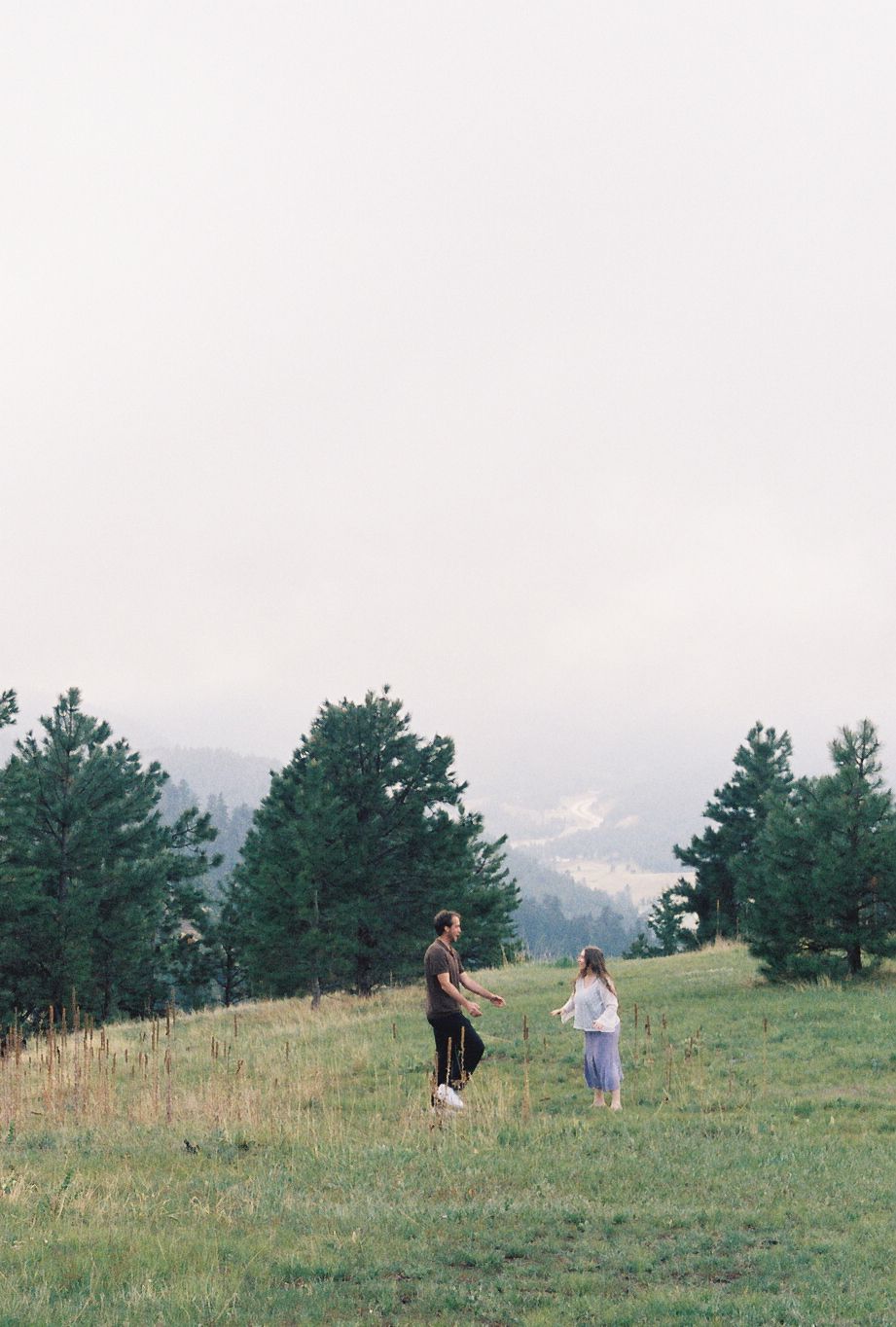 Couple walking together on a lush green hillside meadow surrounded by pine trees on a cloudy day.