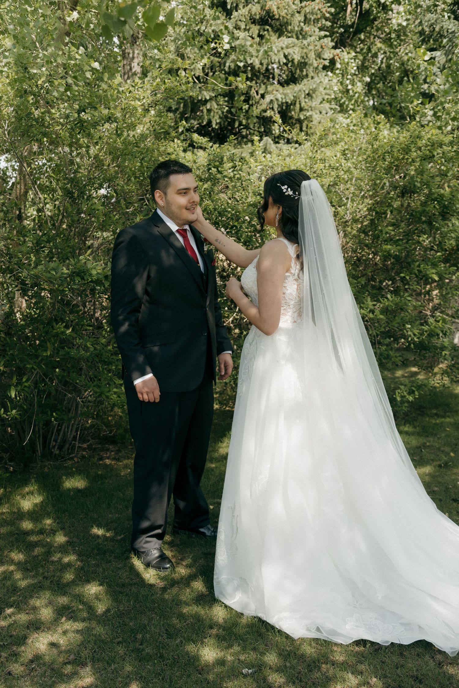 Groom in black suit smiling as bride in white gown touches his face outdoors.