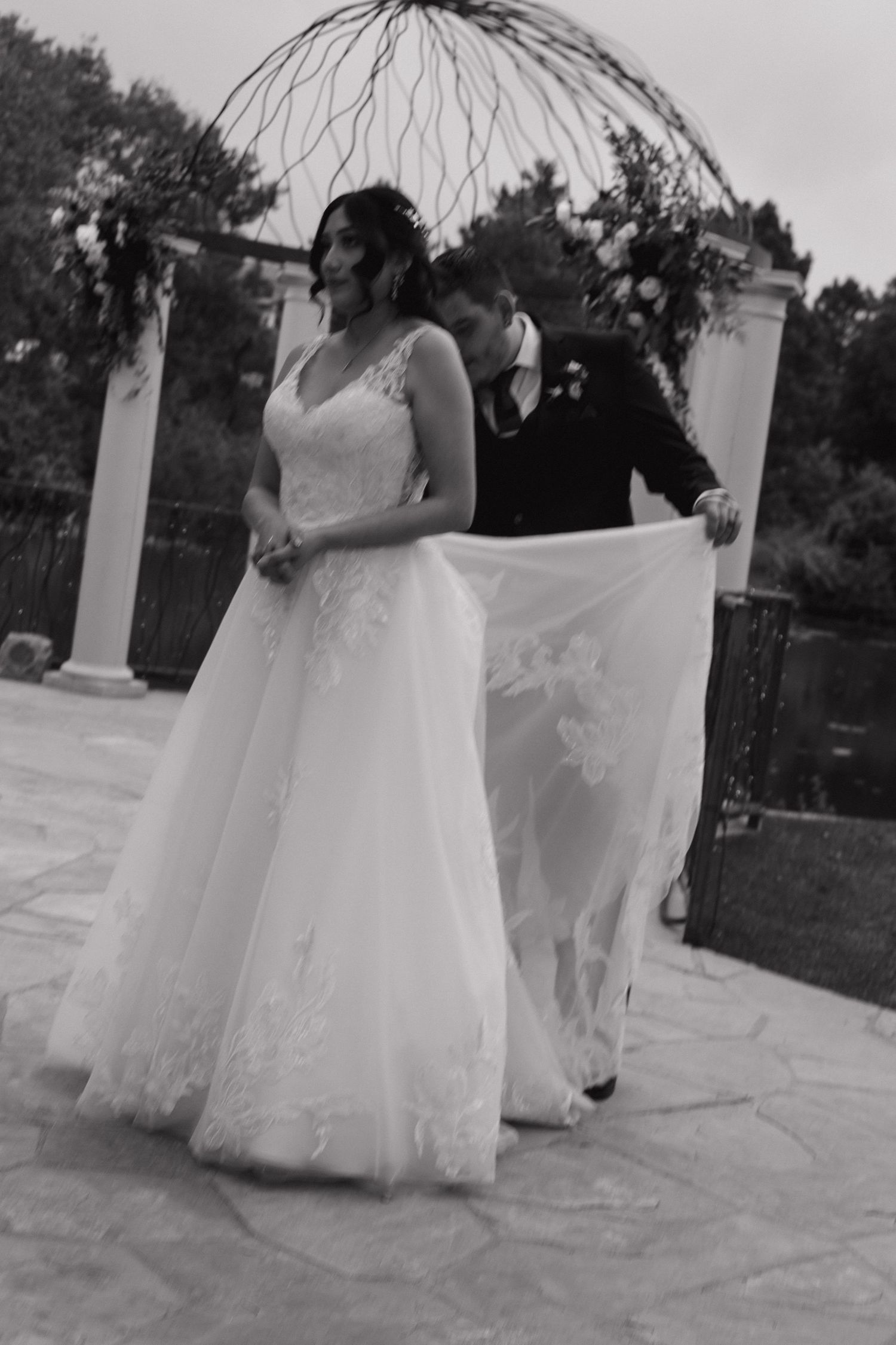 Black and white photo of bride in lace wedding gown with groom adjusting veil at outdoor ceremony altar.