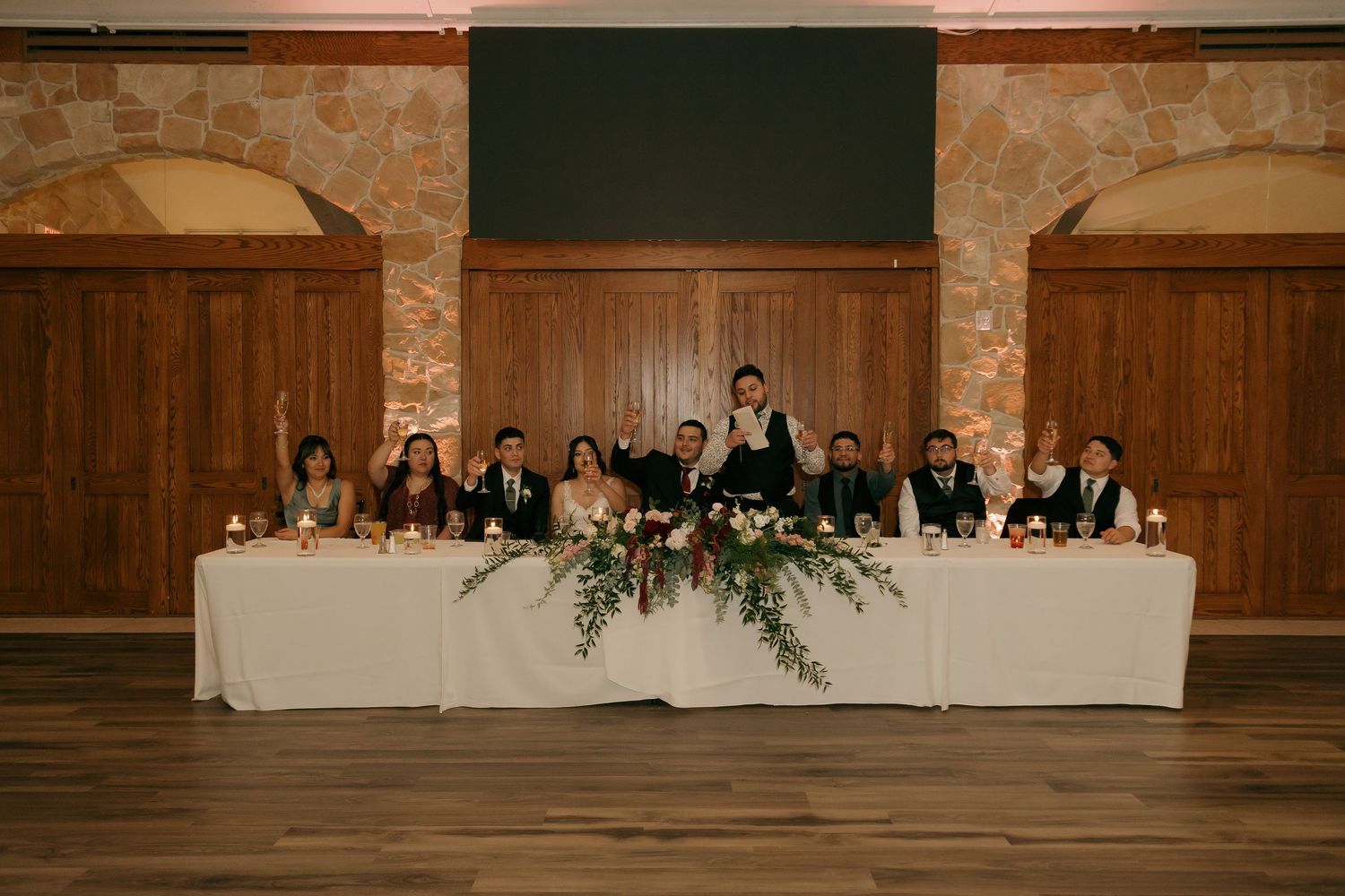 Wedding party seated at decorated head table with floral centerpiece in rustic reception venue.