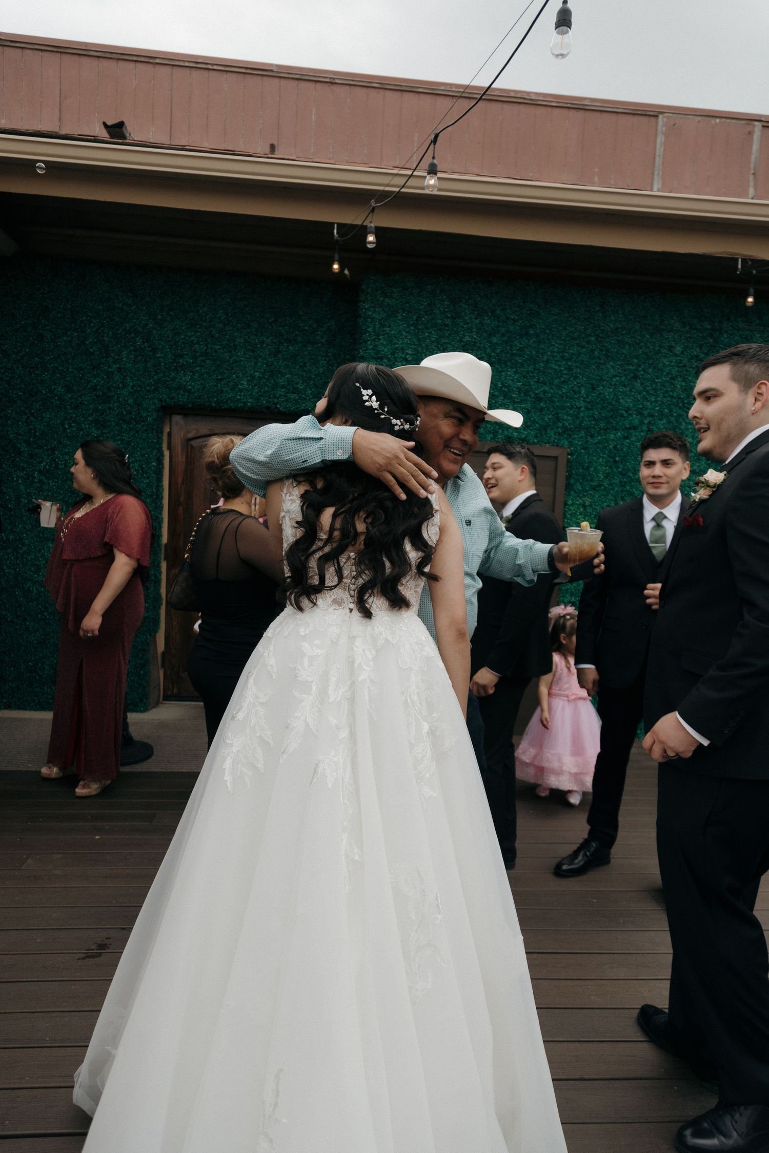 Bride in white gown hugs man in cowboy hat during outdoor wedding reception dance.