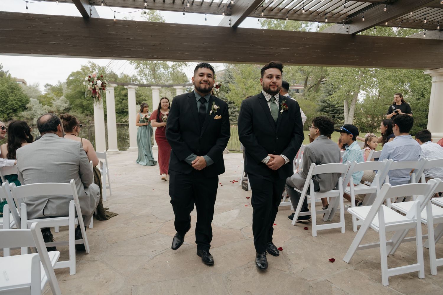 Two groomsmen walk down the wedding aisle at an outdoor ceremony with seated guests.