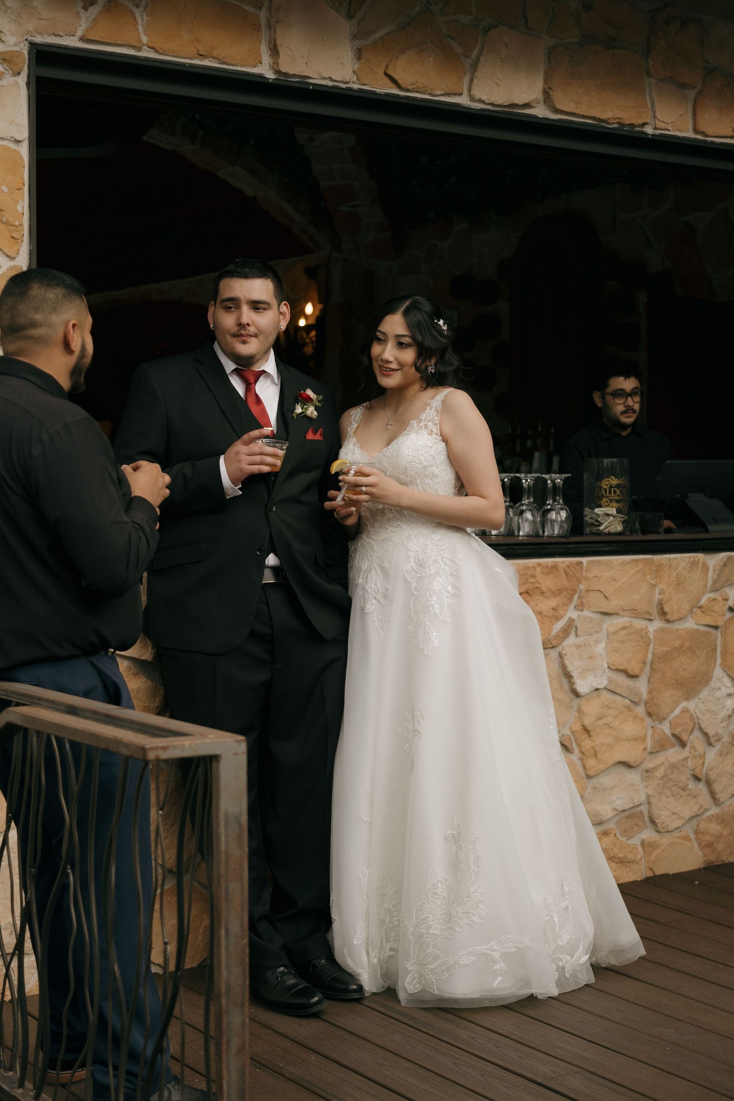 Bride in white lace gown and groom in black suit holding drinks at rustic stone bar.